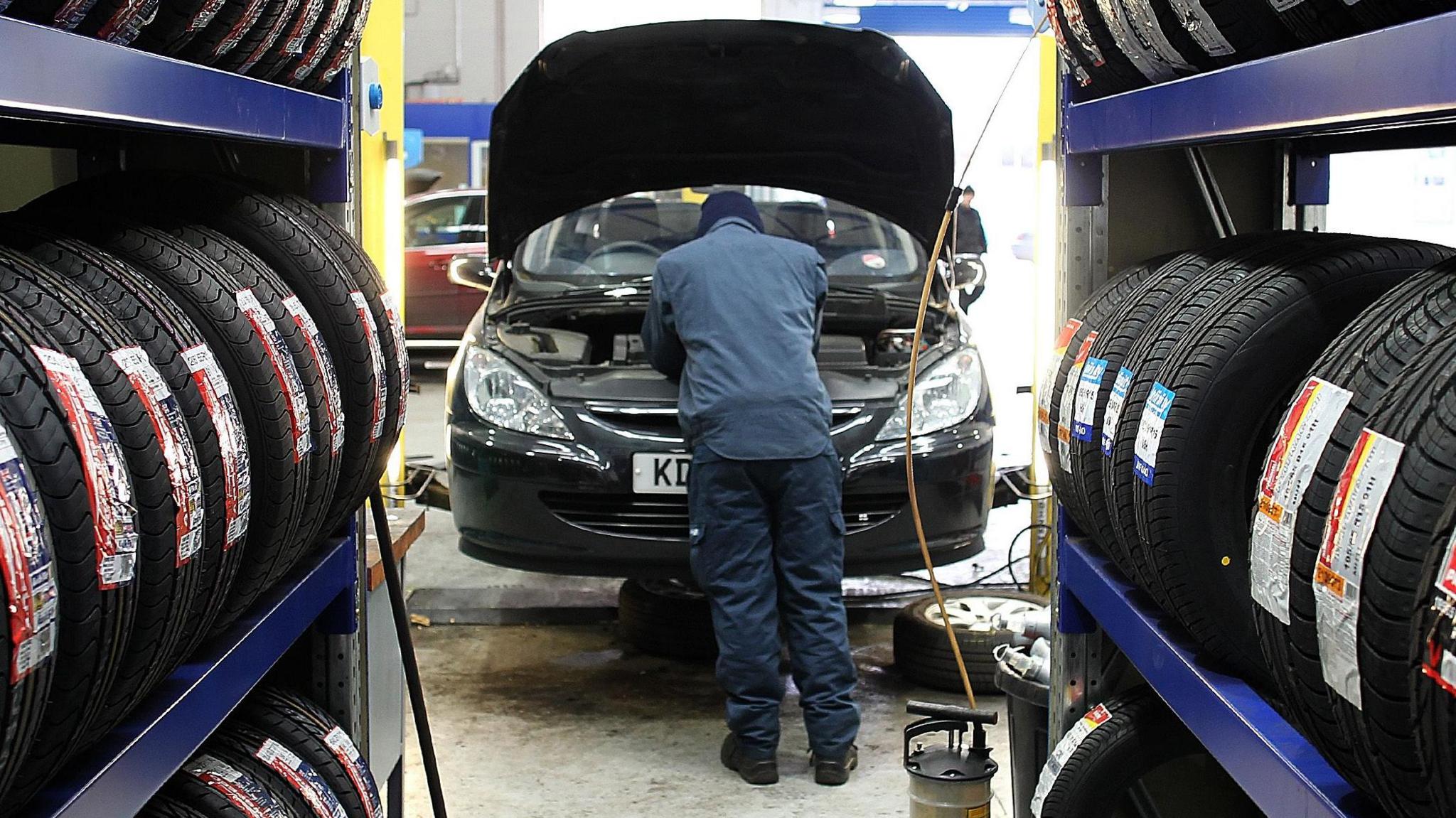 Generic image of a mechanic checking a car's engine under the bonnet inside a garage. Shelves of tyres can be seeen on both sides.