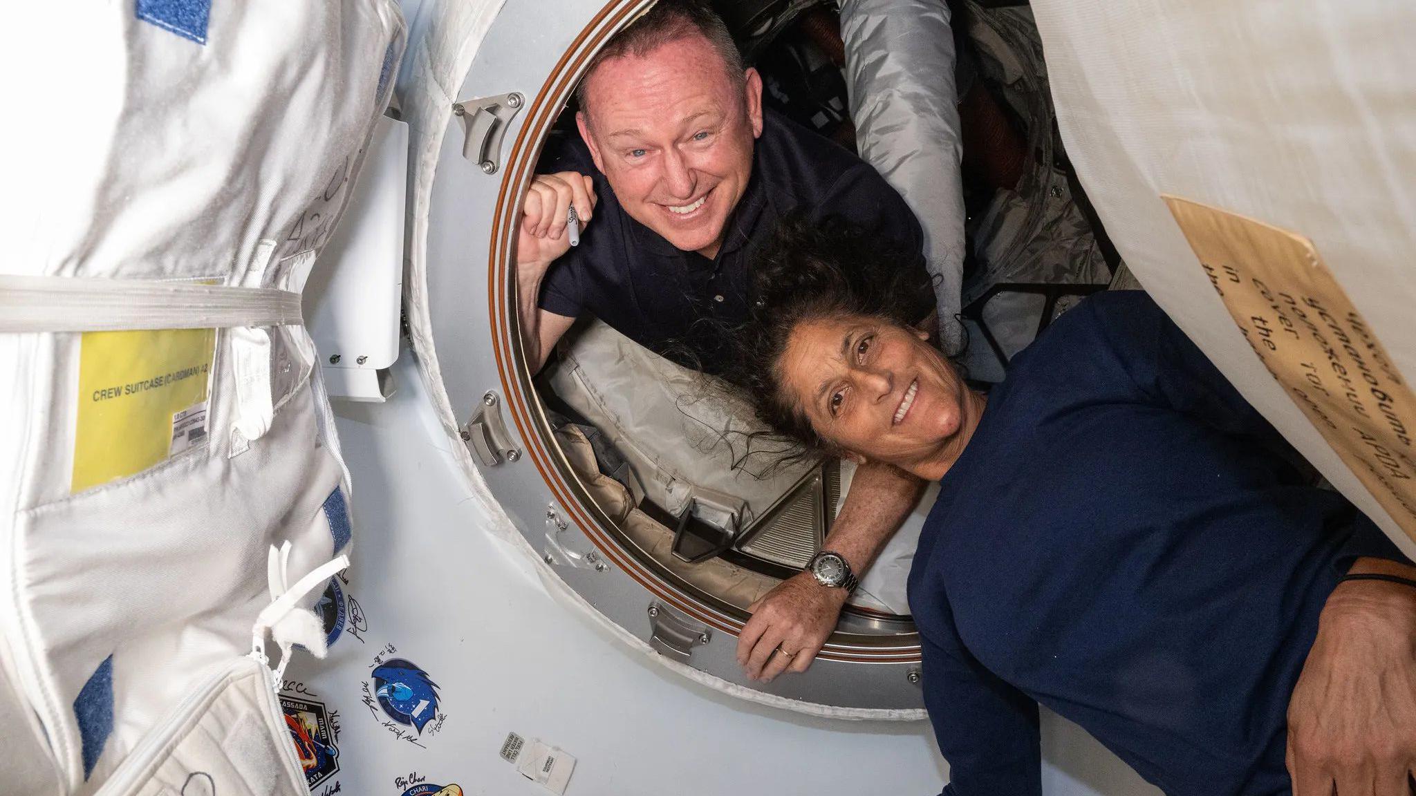 Butch Wilmore looking through a hatch on the international space station and Suni Williams lying just outside. Both are smiling and Suni's hair is waving in the zero gravity environment