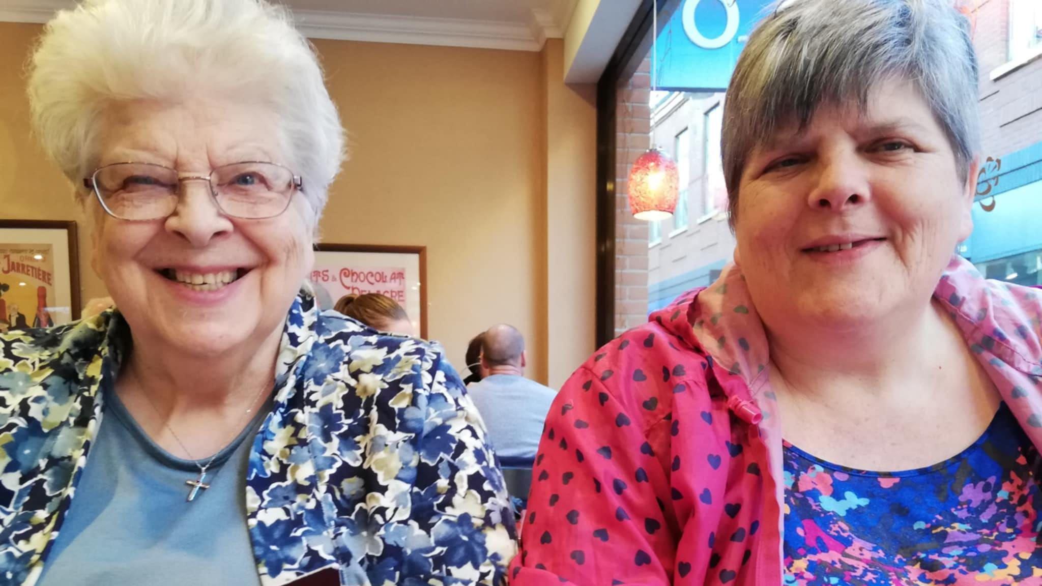 Angela Lennox (on the right) and her mother sitting in a cafe and smiling. Miss Lennox's mother has short white hair and glasses and is wearing a blue jacket with flowers on it. Miss Lennox has short grey hair and is wearing a pink jacket with purple hearts on it.