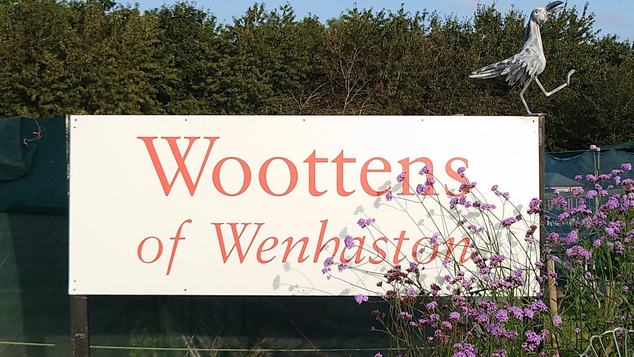 A large white sign with red writing on it that reads Woottens of Wenhaston. Pink flowers are covering part of the sign.