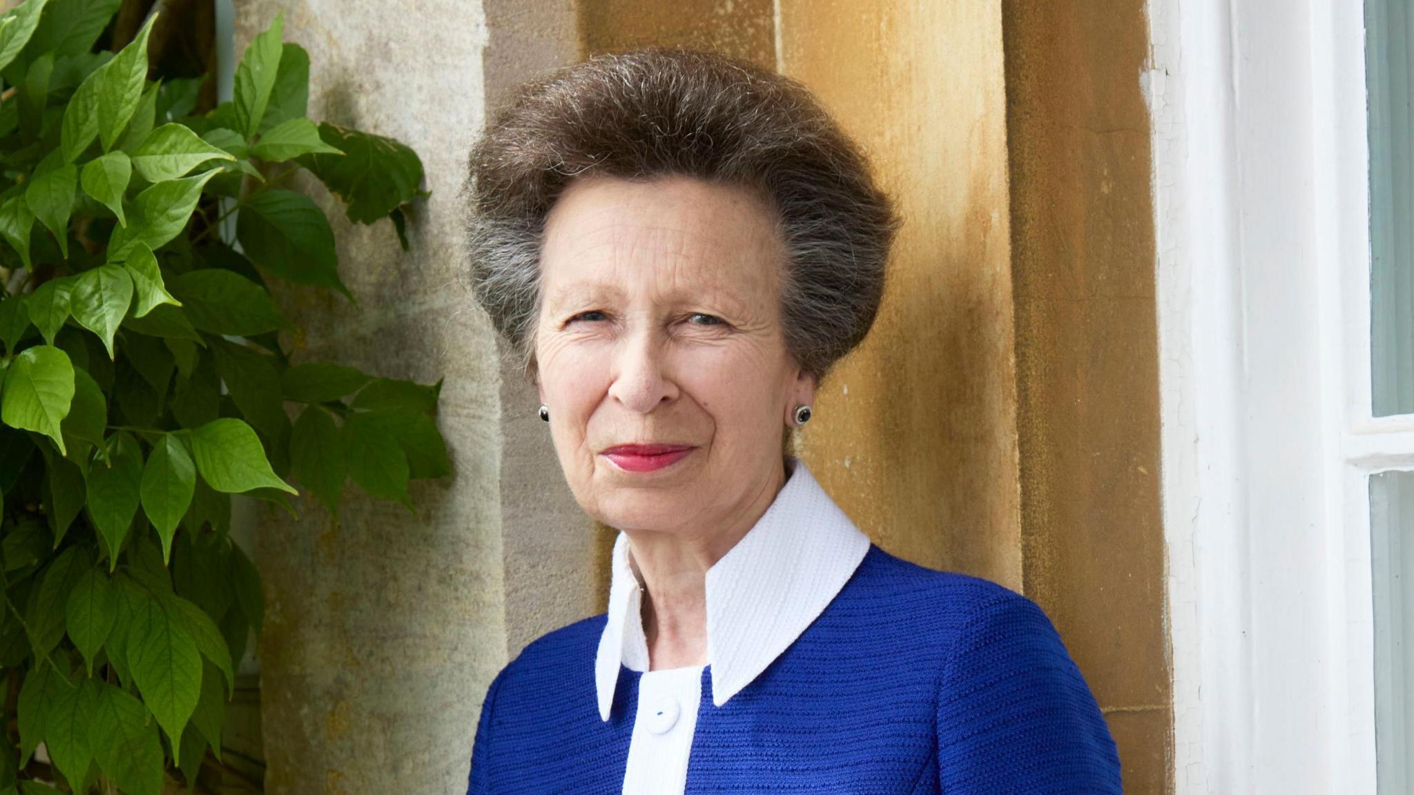 Princess Anne smiles at the camera in a head and shoulders shot. She is wearing a blue jacket with white detail on the front and collar
