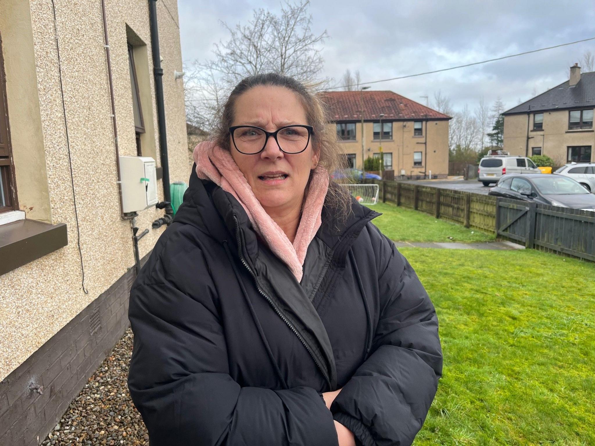 Joy Davidson - a woman wearing black framed glasses with hair tied back, wearing a pink fluffy robe underneath a black padded jacket, standing in the garden in front of her house in Bathgate.