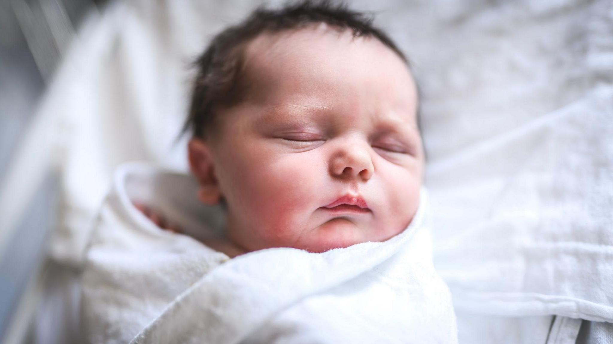 A newborn baby with lots of dark hair lies asleep in a crib