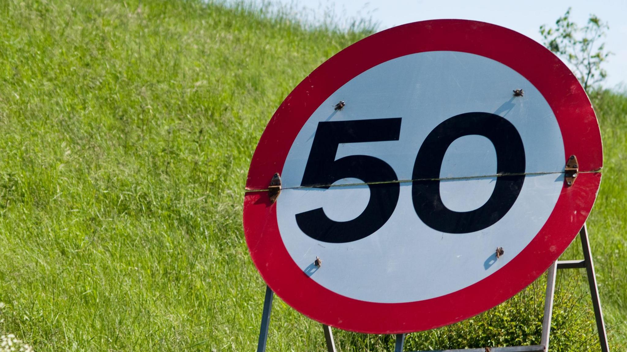 A 50mph sign on a stand in front of a green grass banking