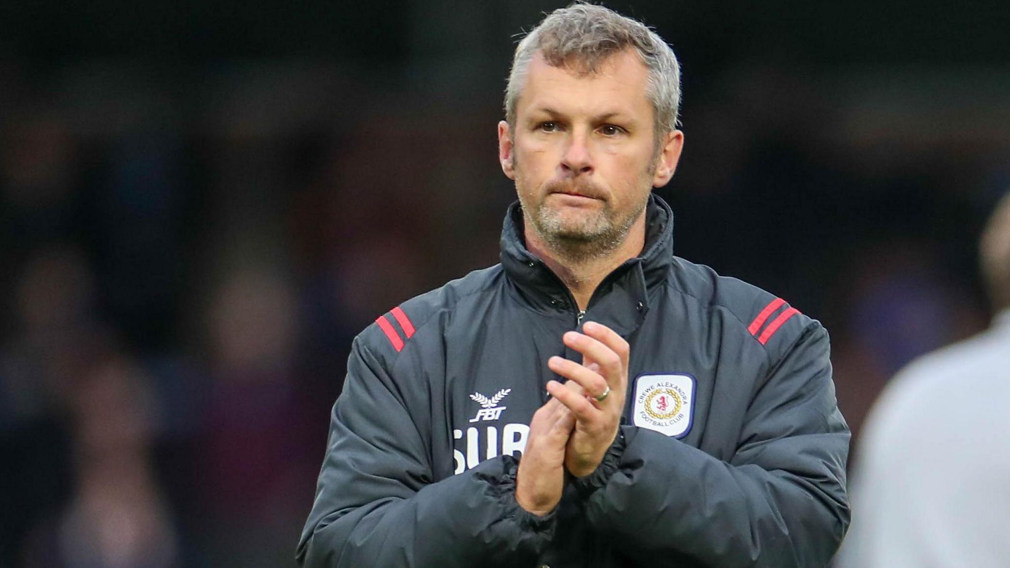 Kenny Lunt applauds the crowd after a game for Crewe. He is wearing a dark grey club coat.