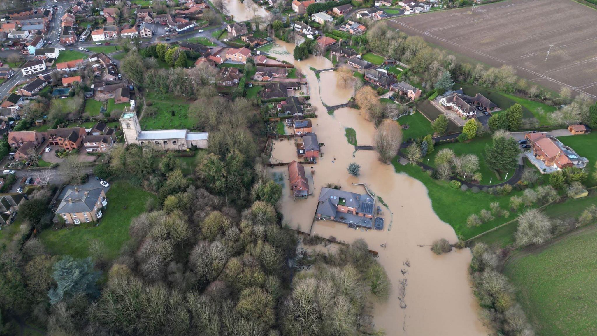 An aerial view of the street, which is partly under water. On the left is housing and a church, surrounded by green land and trees, while on the right is more housing and a stretch of farmland 