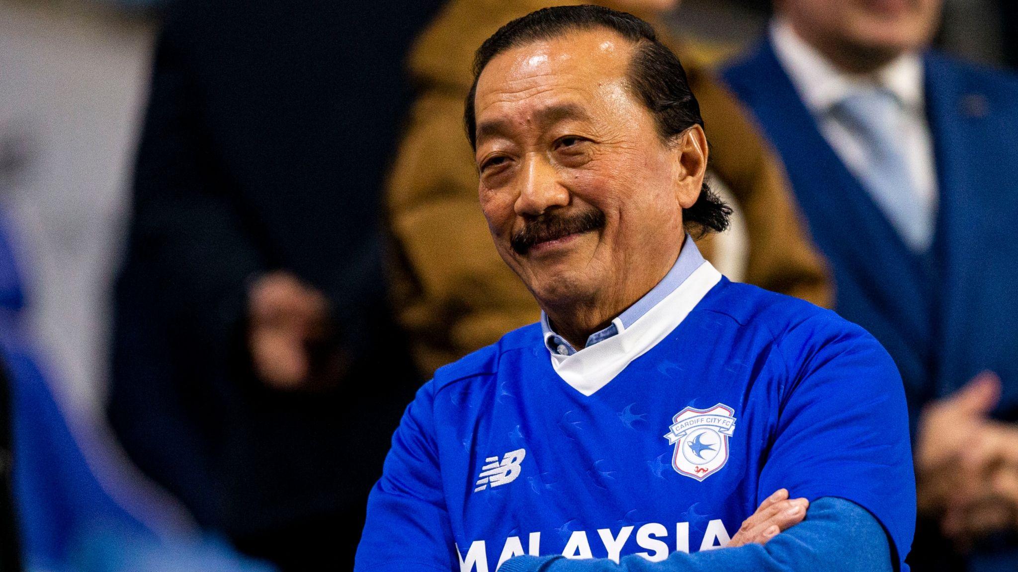 Vincent Tan in a blue Cardiff City shirt smiles with arms folded in the stands at a Cardiff City games