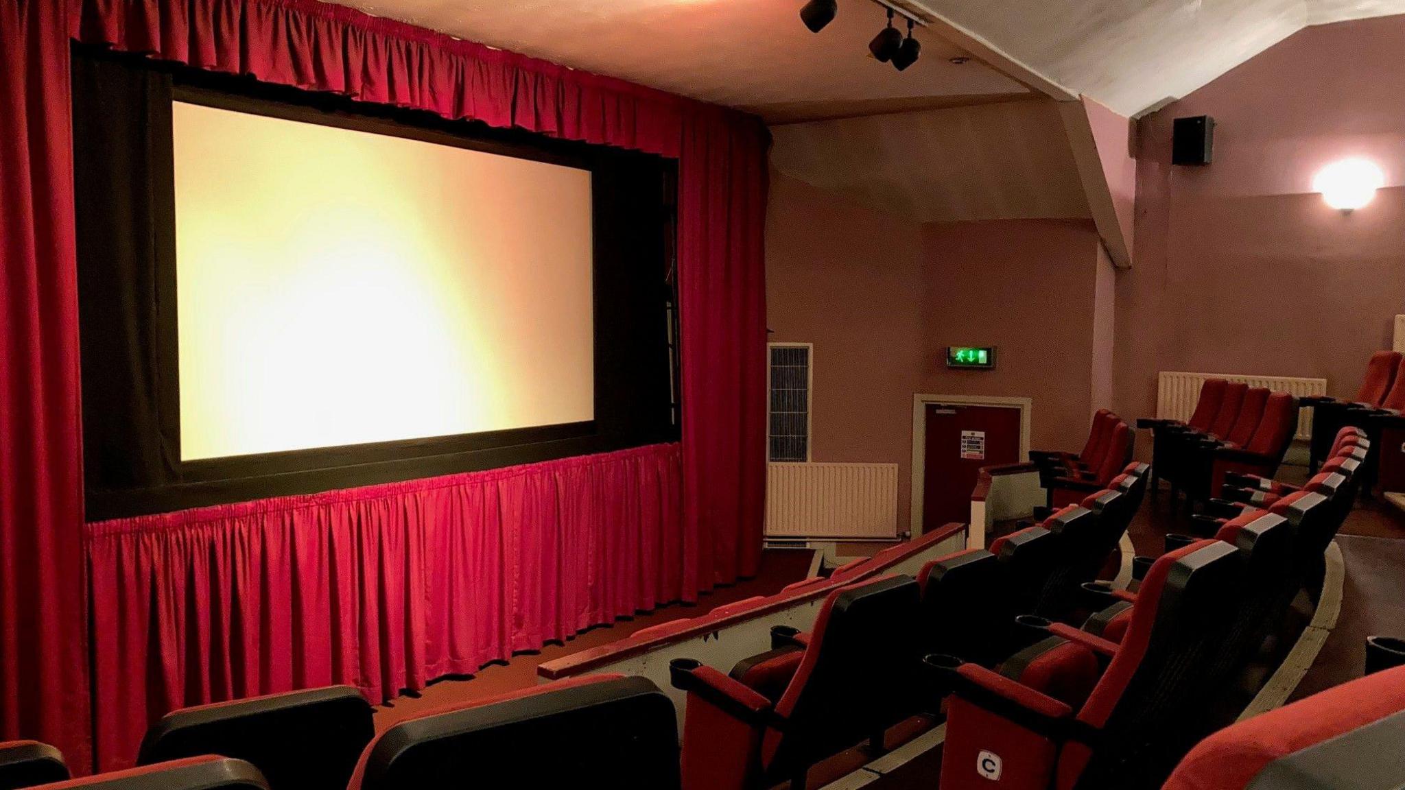 A cinema screen fringed with a red curtain and red velvet seats in a semi-circle facing towards it.