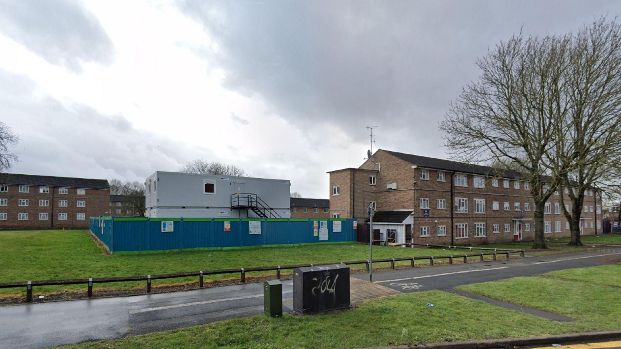 Brown brick blocks of flats surround a green space, on which sits a grey temporary building, surrounded by blue fencing.