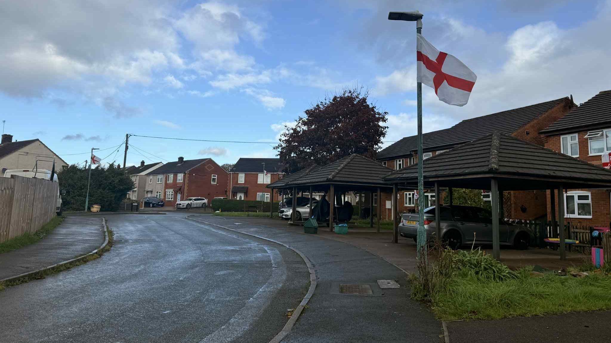 A St George's flag flies in the wind fixed to a lamppost on the inside edge of a curved pavement. Along the right side of the image there are orange bricked houses and cars parked on driveways. There is a bright blue sky blotted with dark grey clouds.