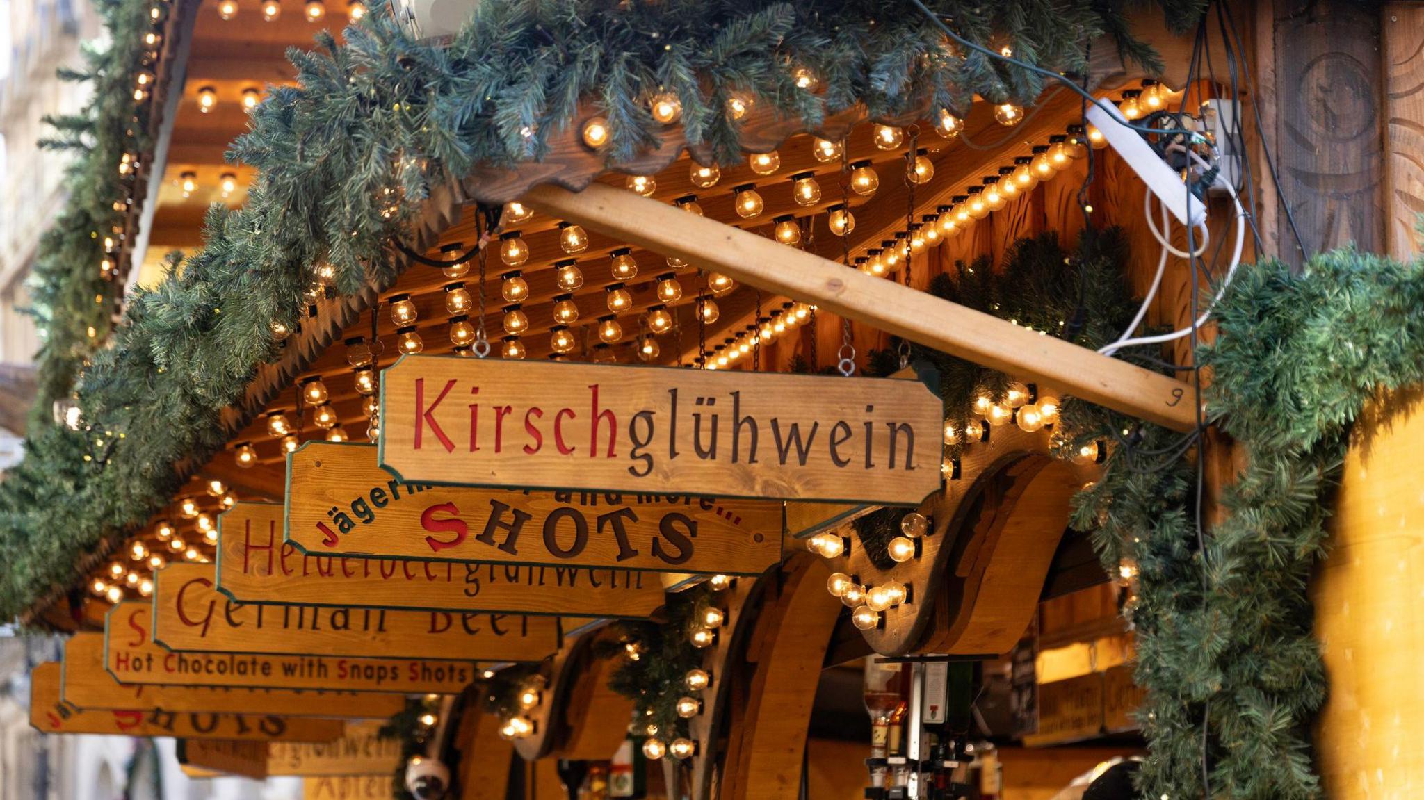 Wooden signs lined up over a wooden stall, in German , advertising wares such as Kirschgluhwein, shots and German beer. The stalls are adorned with fir tree garlands and have light garlands strung up.