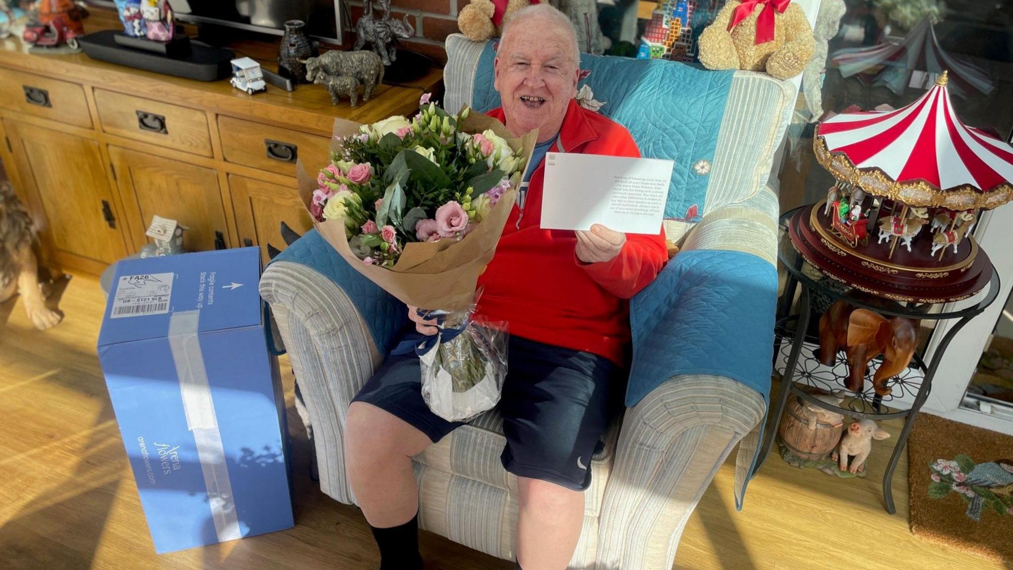 A man wearing a red top and shorts, sits on a chair while holding a bouquet of flowers in one hand and a note in the other.