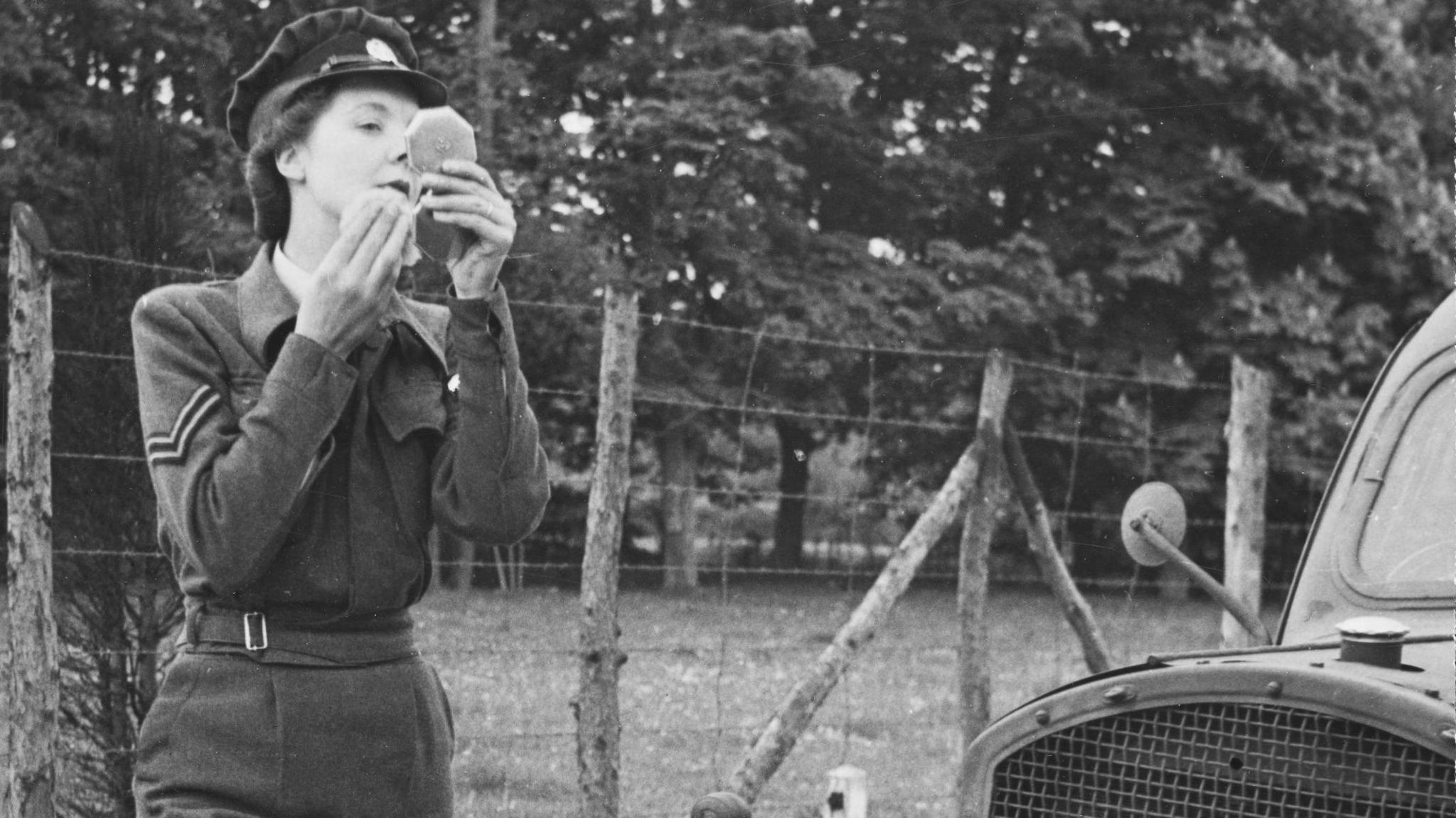 Peggy Harvey, in her uniform, doing her makeup, while standing next to a military vehicle. There is a wire fence with wooden posts behind her. In the background is trees.