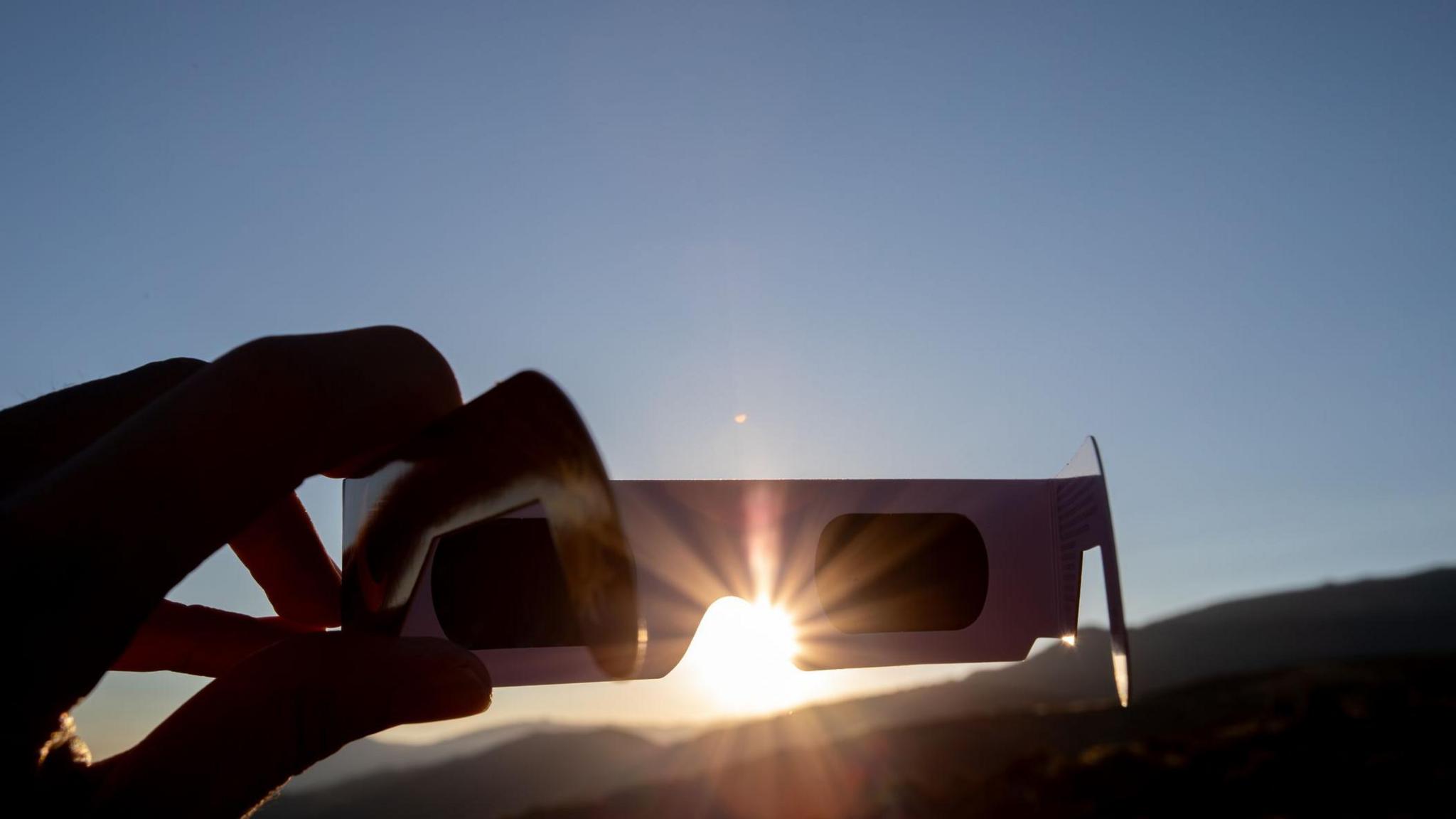 Person holding eclipse glasses up to the sun with sunbeams shining through