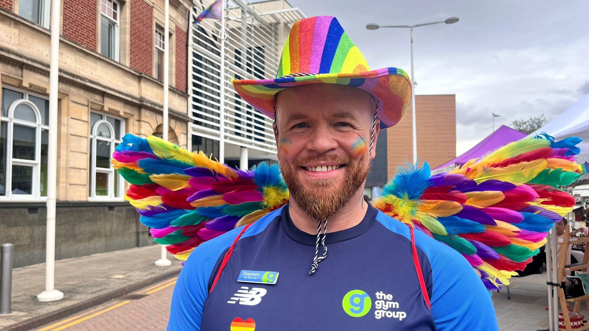 A man with a reddish-brown beard smiles at the camera. He is wearing a rainbow cowboy hat and rainbow coloured angel wings.