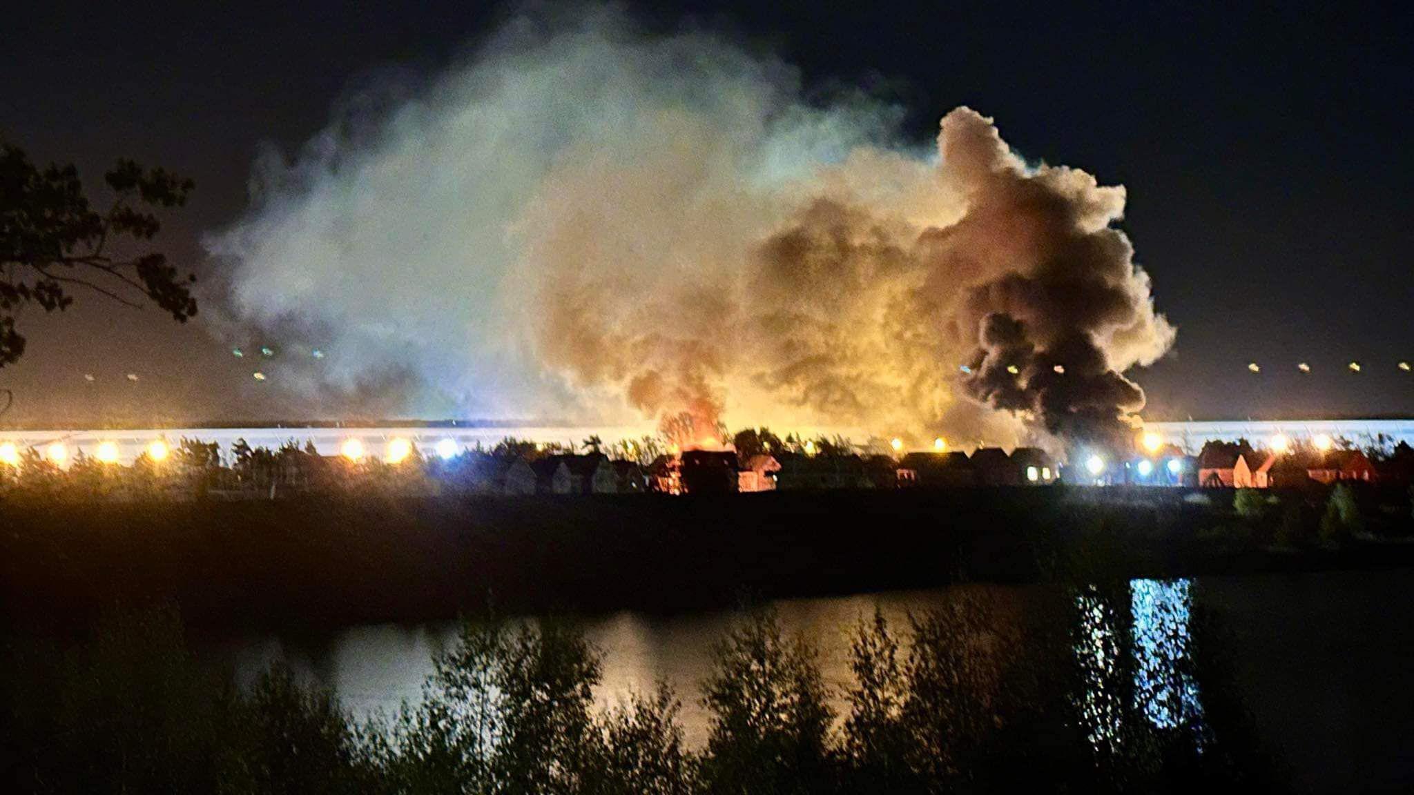 A night image of a row of houses on fire with large white clouds of smoke above. The properties are next to a lake or body of water.