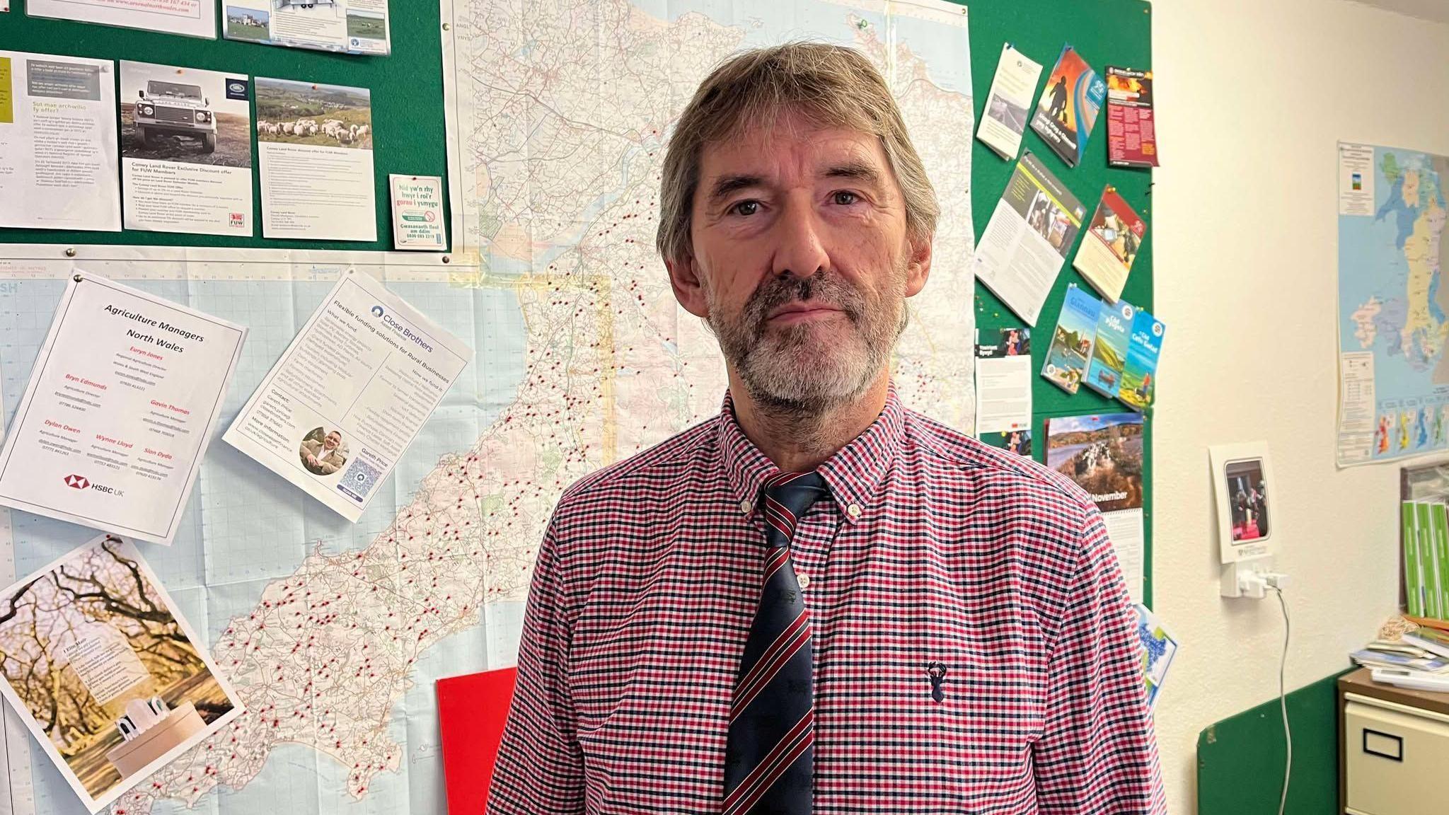 Gwynedd Watkin, county officer of the Welsh Farmers' Union in Gwynedd, within an office setting, he stands in front of a large pin board covered in maps and leaflets. He has a grey and brown speckled beard, and floppy hair. He wears a red, navy and white small checked shirt and wears a navy and red horizontal striped tie. 