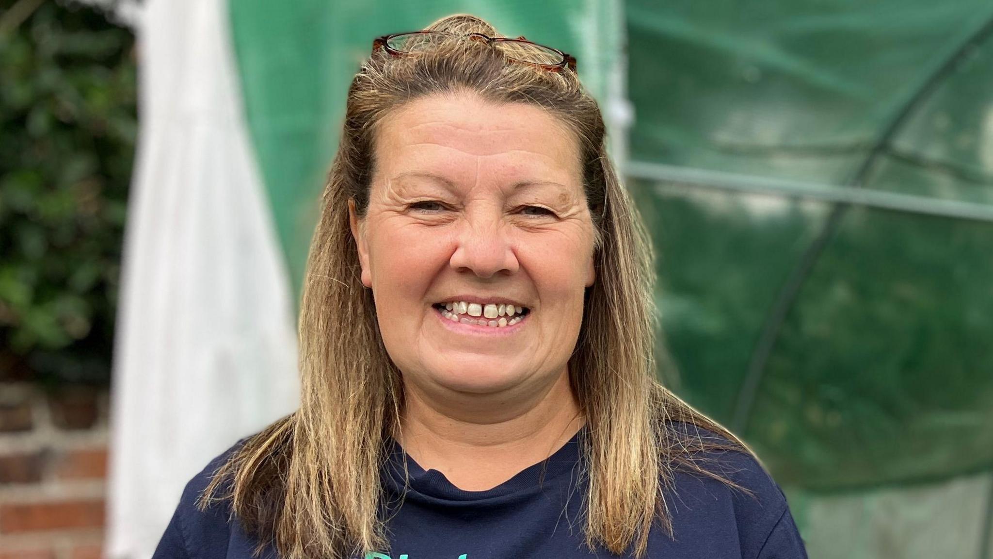 Penny standing in front of a polytunnel with green netting. She is smiling at the camera. She has a navy blue T shirt and shoulder length light brown hair, with glasses aloft her head.