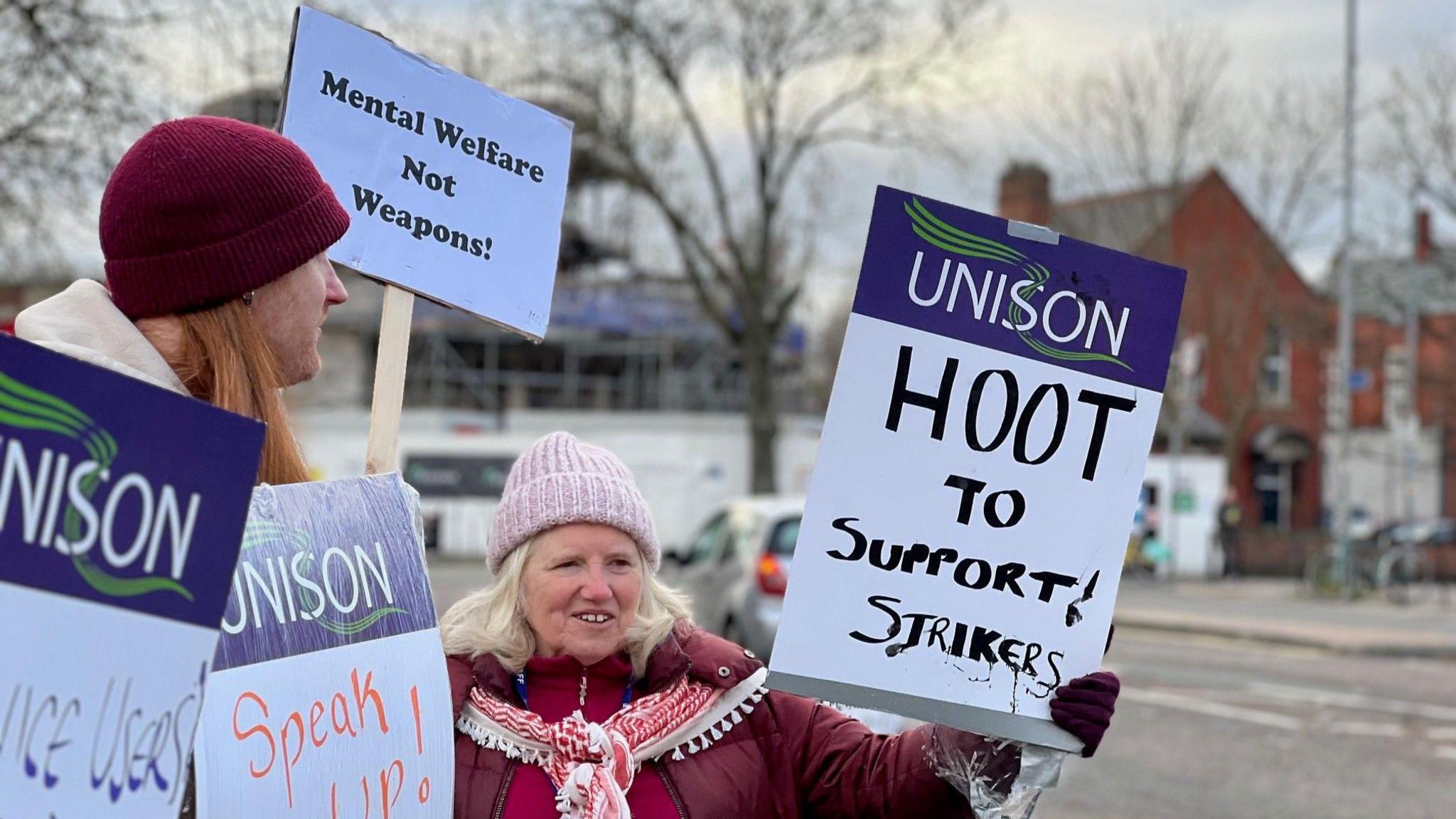 Two NHS workers strike, holding Unison union placards, with one saying 'hoot to support strikers' and another saying 'speak up!' A woman is wearing a pink woolly hat and is wearing a burgundy coat with a red and white scarf. A man standing next to her is wearing a burgundy woolly hat and has long red hair. Some buildings can be seen across the side of the road from where they are standing.