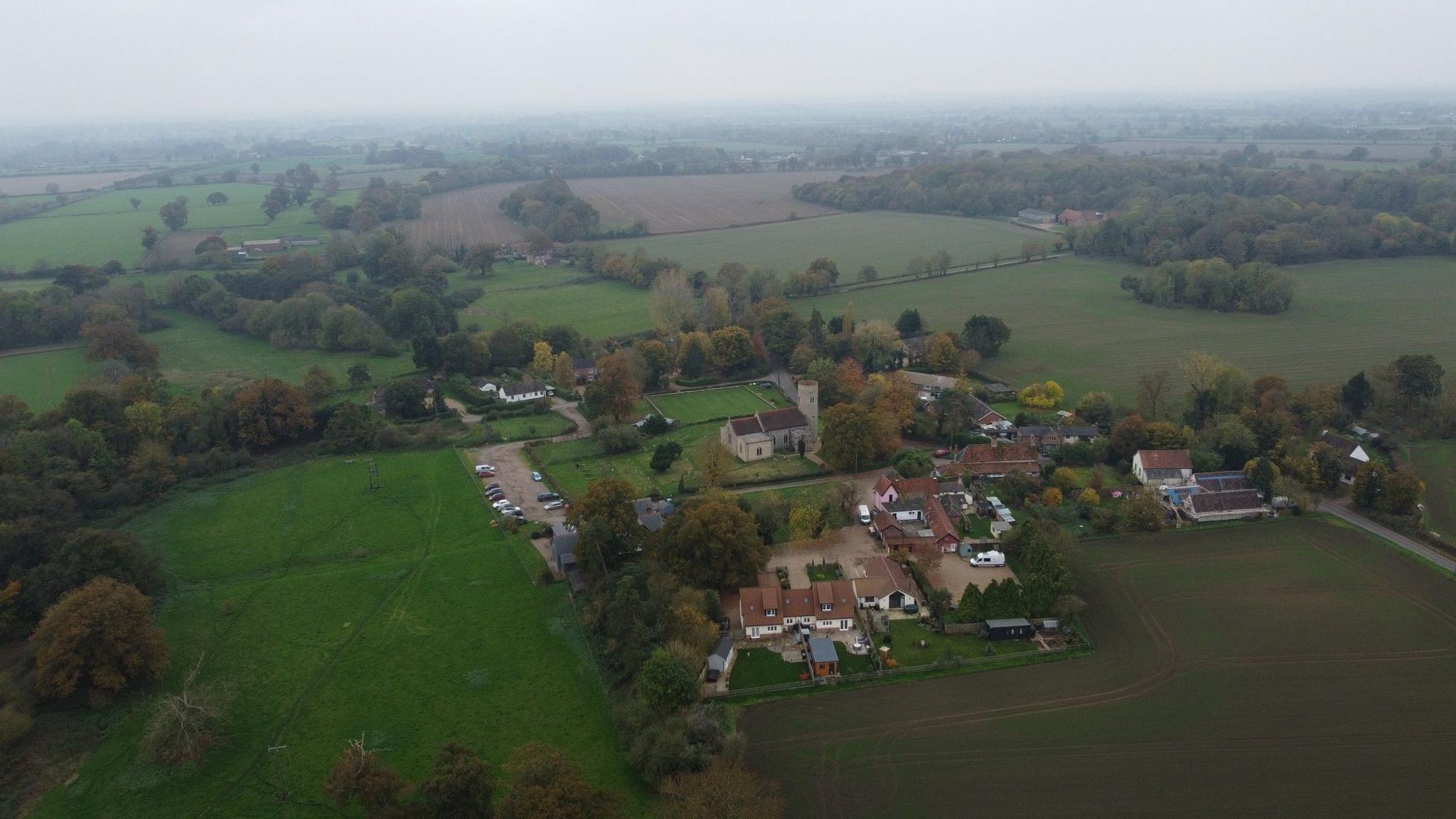 Aerial view of the village of Gissing, surrounded by fields and a large cluster of farm buildings including some houses and farm sheds as well as the parish church.