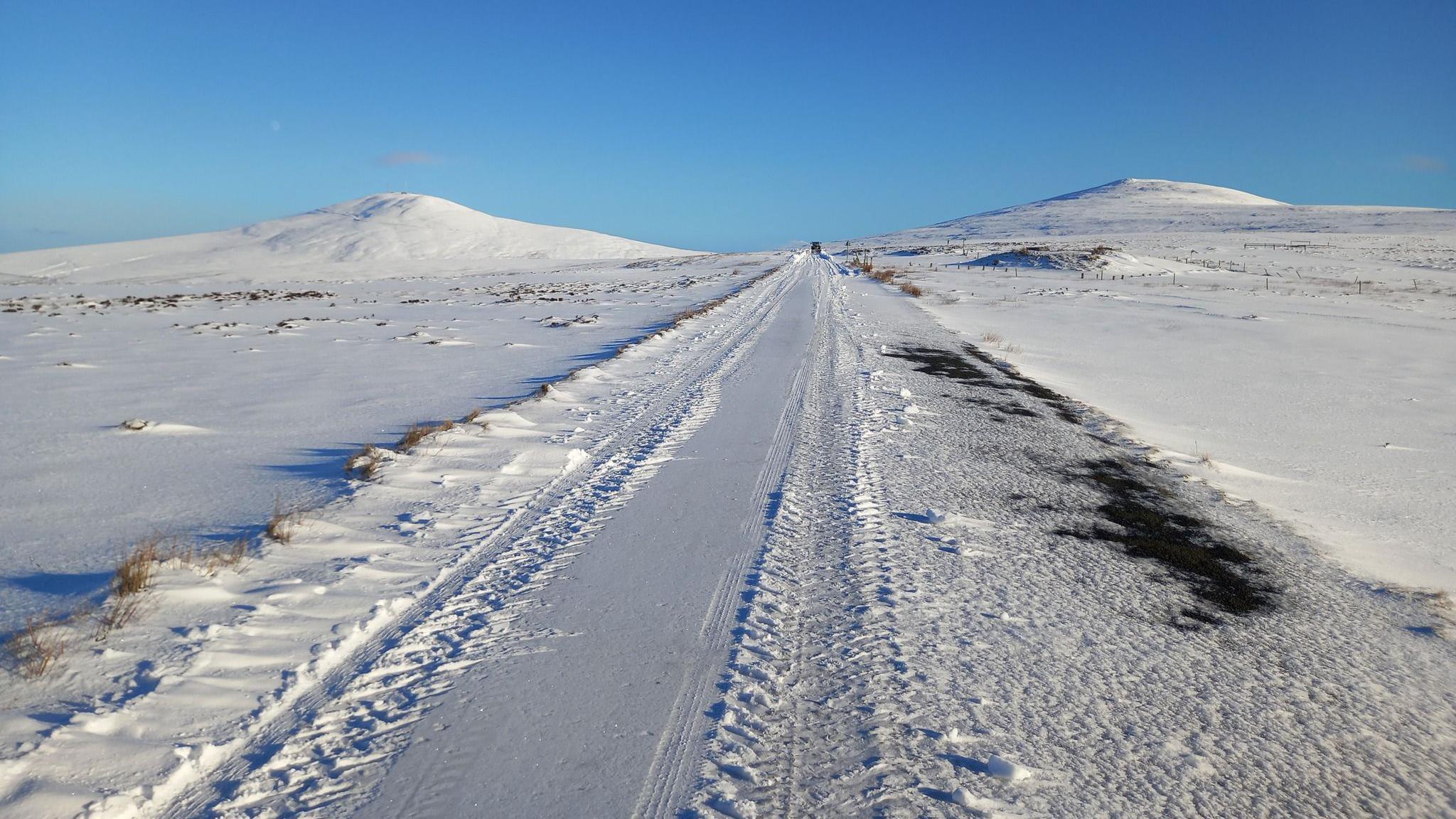 A road covered in snow, between two peaks of hills.