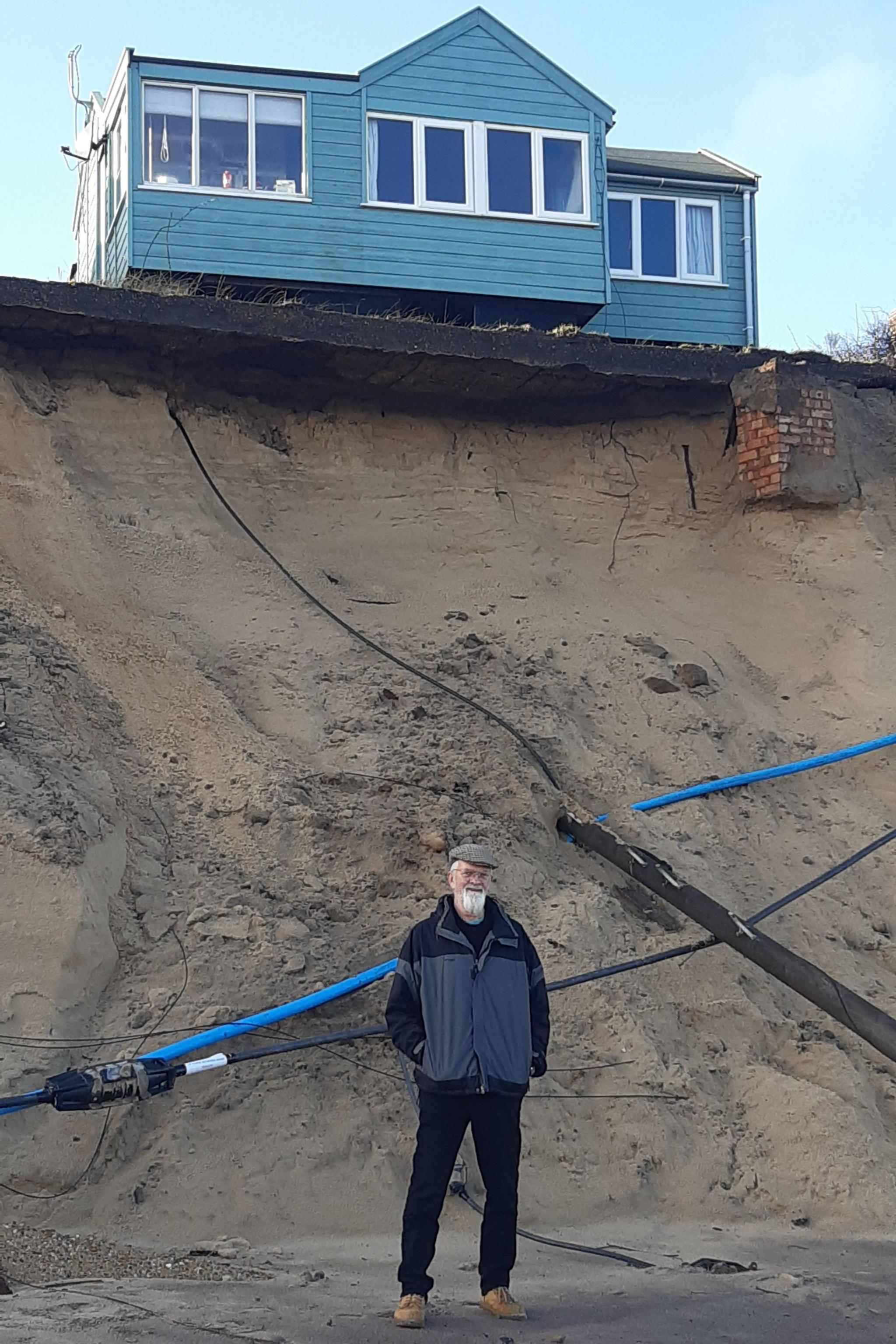 Andy Sloan wearing a hat and standing three or four metres below his chalet, which is perched on the edge of a road left exposed by coastal erosion.