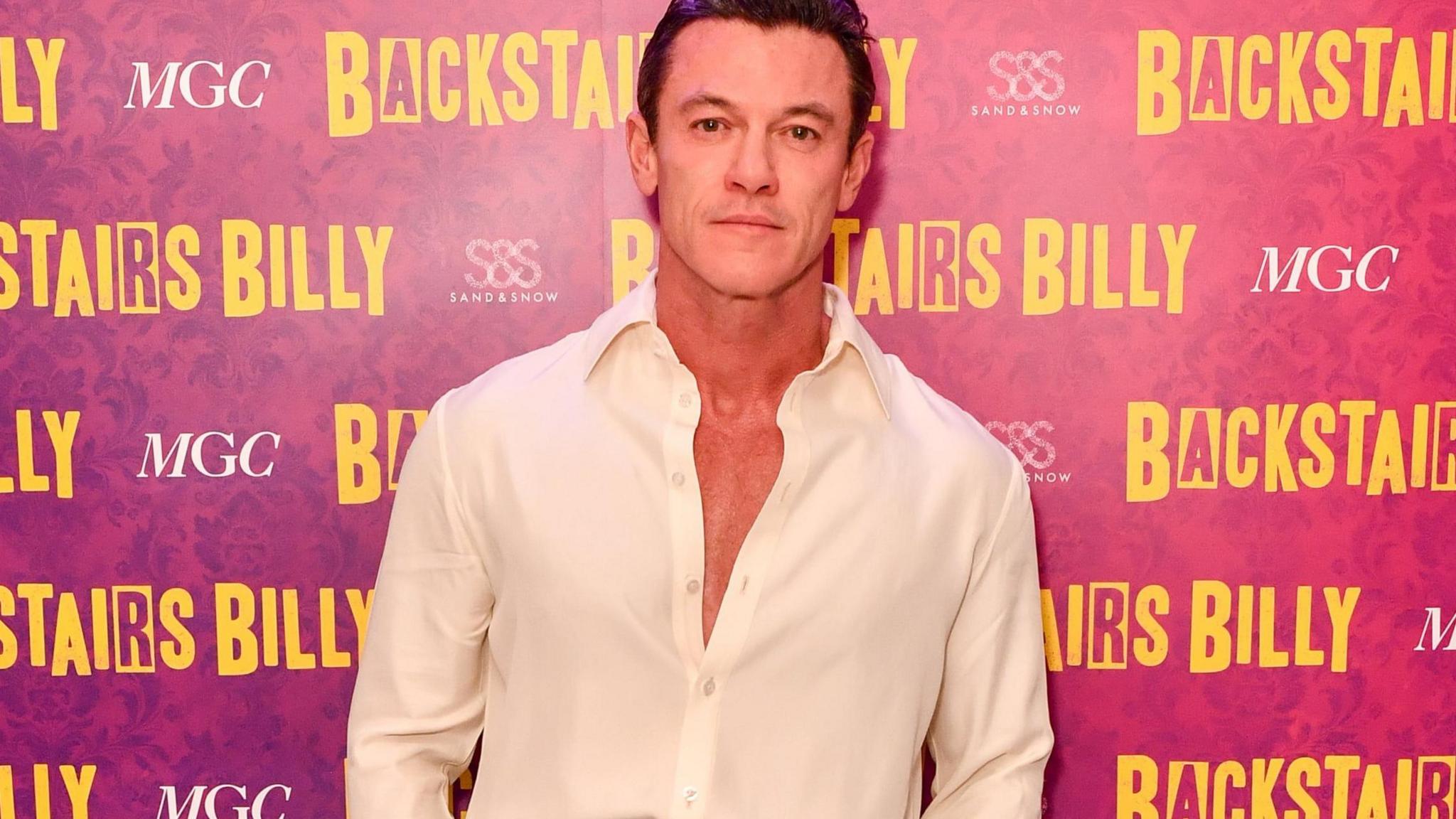 Welsh actor Luke Evans standing in front of a background that says Backstairs Billy, he is wearing a whit shirt that is unbuttoned. 