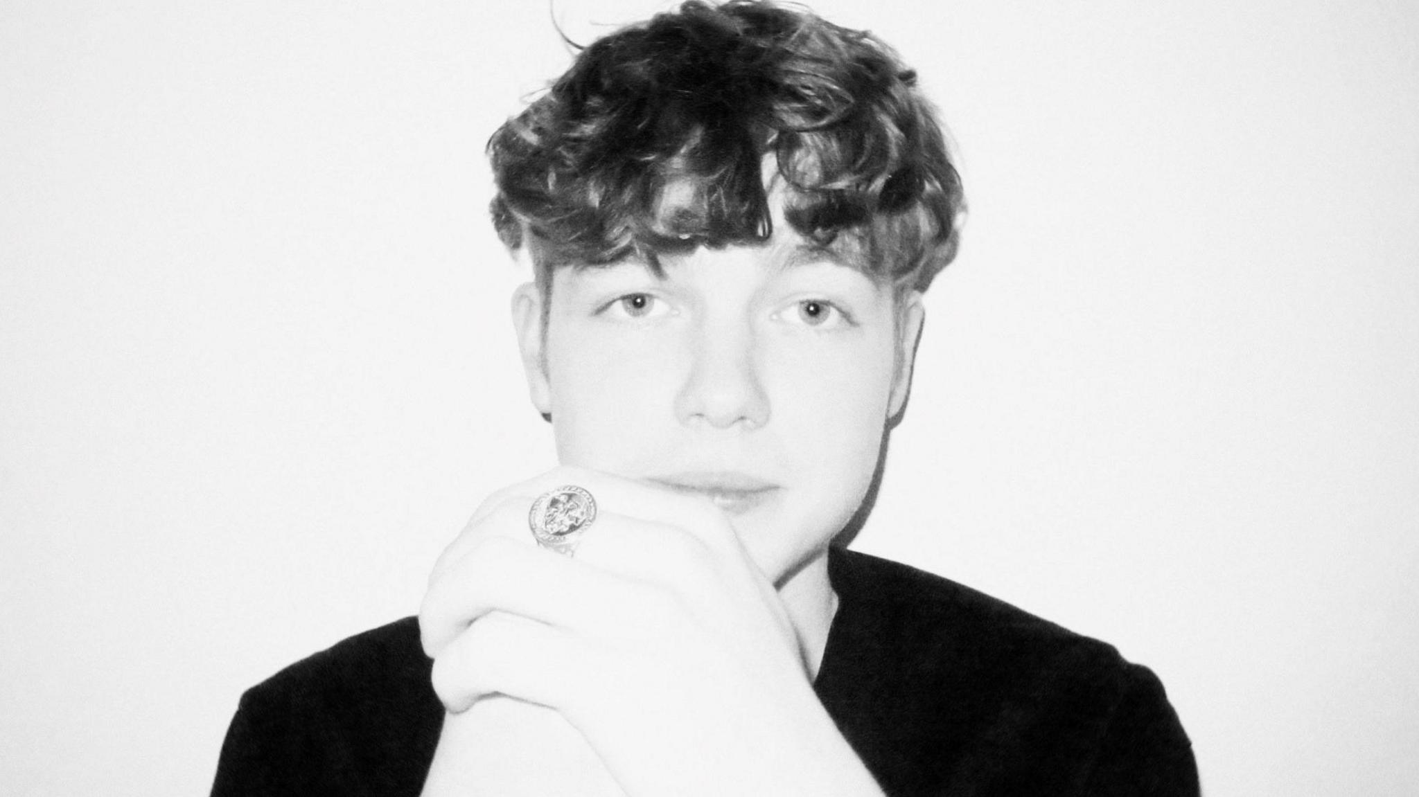 DJ Shelly. Shelly is an 18-year-old white man with short curly hair. In this black and white photograph, he looks at the camera, his hands folded in front of his face. He wears a large ring on his middle finger and a plain black T-shirt. 