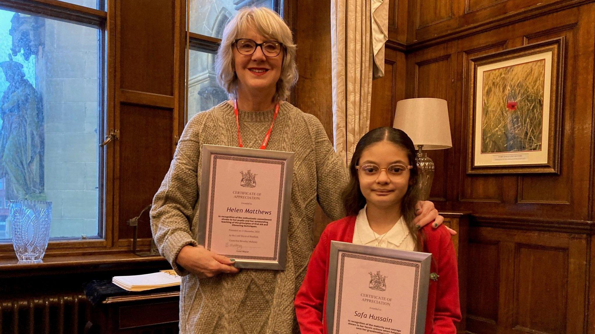 A white-haired woman wearing glasses and grey jumper dress holding a framed certificate standing alongside a young girl with dark, long hair and glasses also holding a framed certificate. 