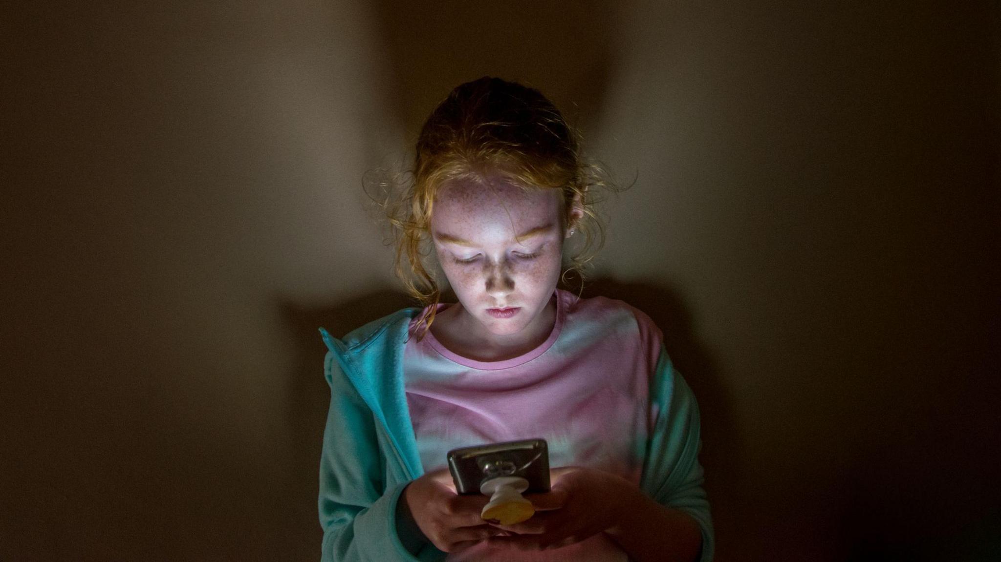 A girl sits in a dark room on her phone. The light from the screen lights up her face. She has a neutral expression.