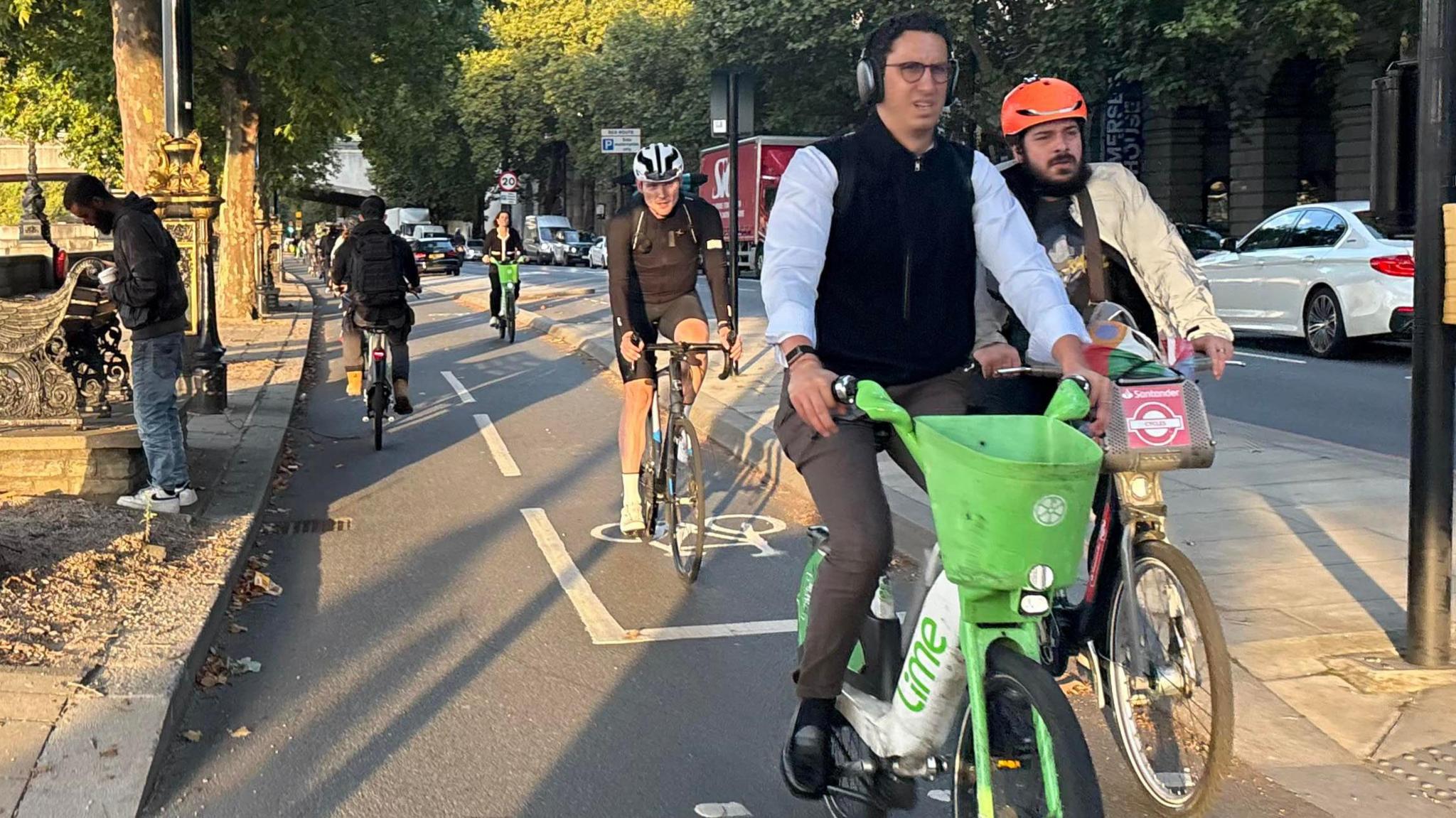 People cycling on a London street. A variety of bikes are used, including lime green Lime E-bikes, red branded Santander E-bikes and non-electric bikes such as drop handlebar road bikes and all-purpose mountain bikes.