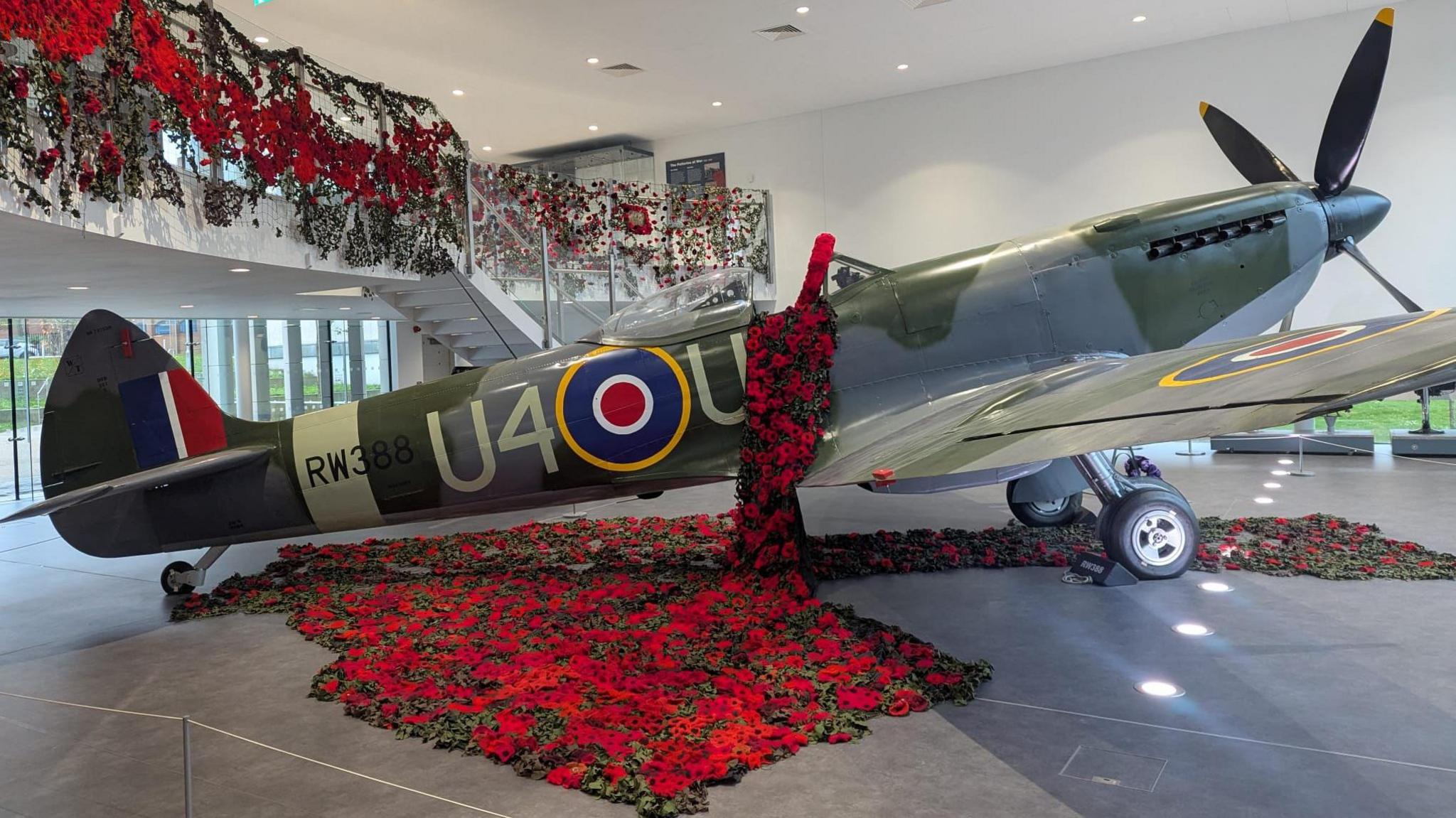 A World War Two Spitfire aircraft, which is a green and grey plane with numbers written on its side, in a room, covered in knitted poppies. Poppies are also hanging from a railing above the plane.
