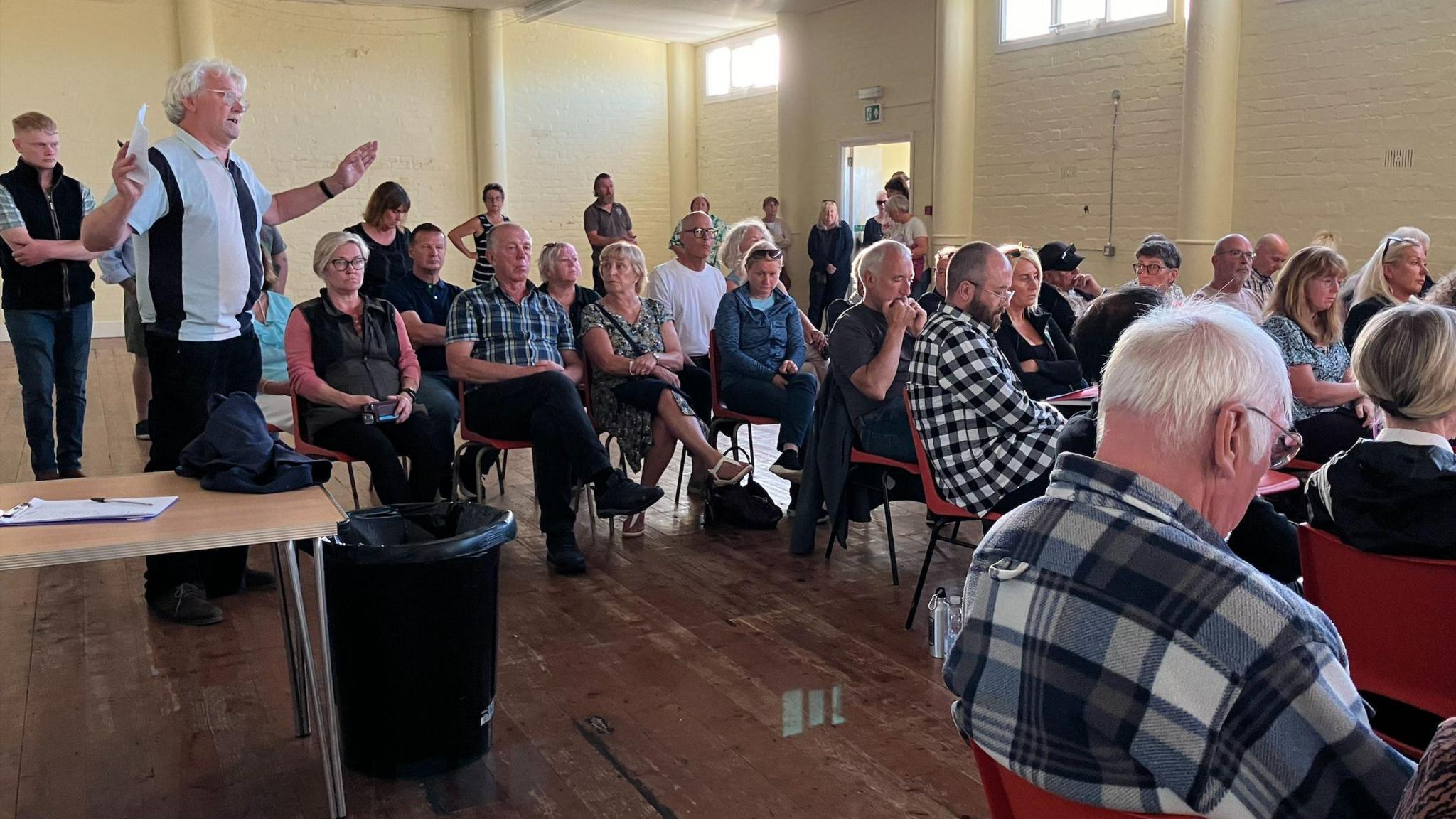 Members of the public in Jurby Parish Hall. About 30 people sit on orange chairs in a hall with yellow walls, with some people standing up behind them. One man in a striped top is standing up gesticulating while asking a question.