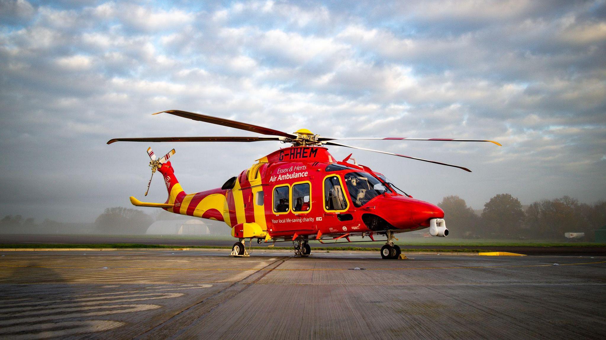 A red and yellow helicopter on an airstrip with the registration G-HHEM and the name of Essex and Herts Air Ambulance on its side.