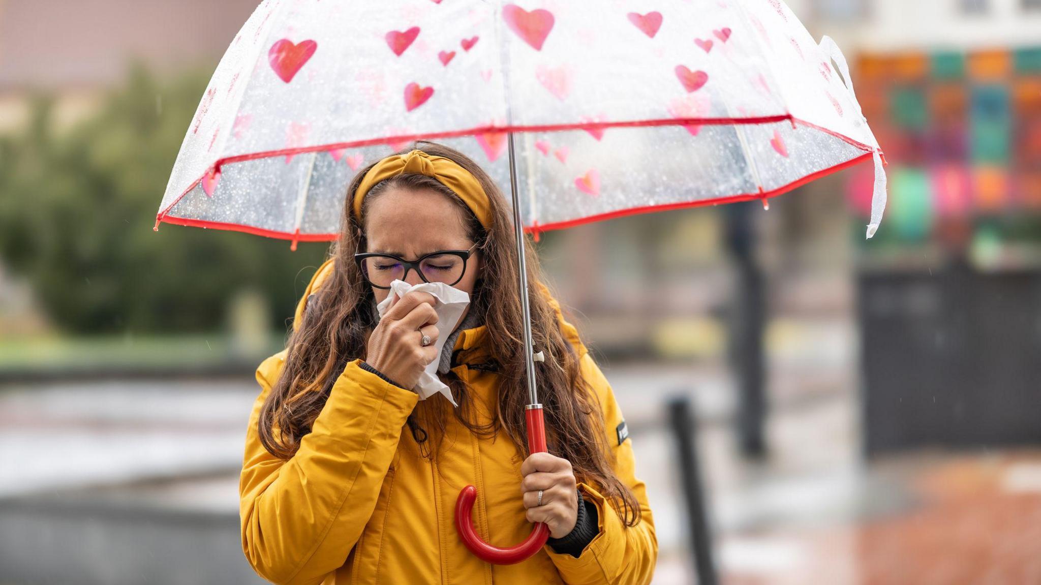 A woman blowing her nose and sneezing into a handkerchief during a rainy autumn day. She is wearing a yellow rain coat and a red umbrella 