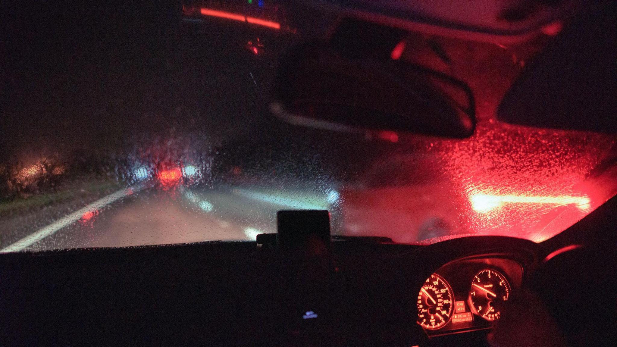 The driver's view out of the front of a car on a dark, rainy night. Inside the car dials on the dashboard are lit up and the rear-view mirror is visible. Through the windscreen you can see blurred oncoming headlights in the distance.