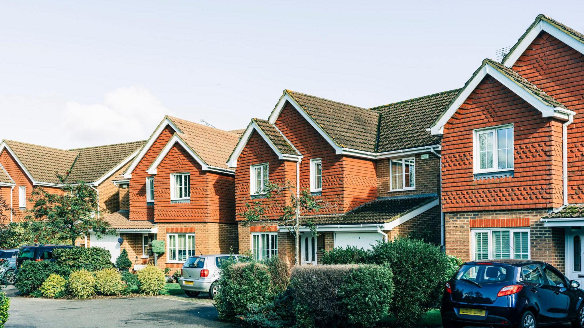 Three residential red brick houses, two stories with large windows at the top and bottom. Cars, silver and blue are parked in leafy driveways. The sky is blue grey and the sun is shining on the houses. 