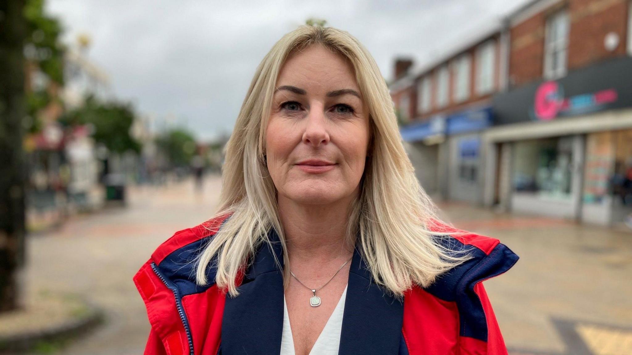 Paula Gouldthorpe has long blonde hair and stands on a shopping street in Scunthorpe, she is wearing a white v neck top and a blue and red coat. 