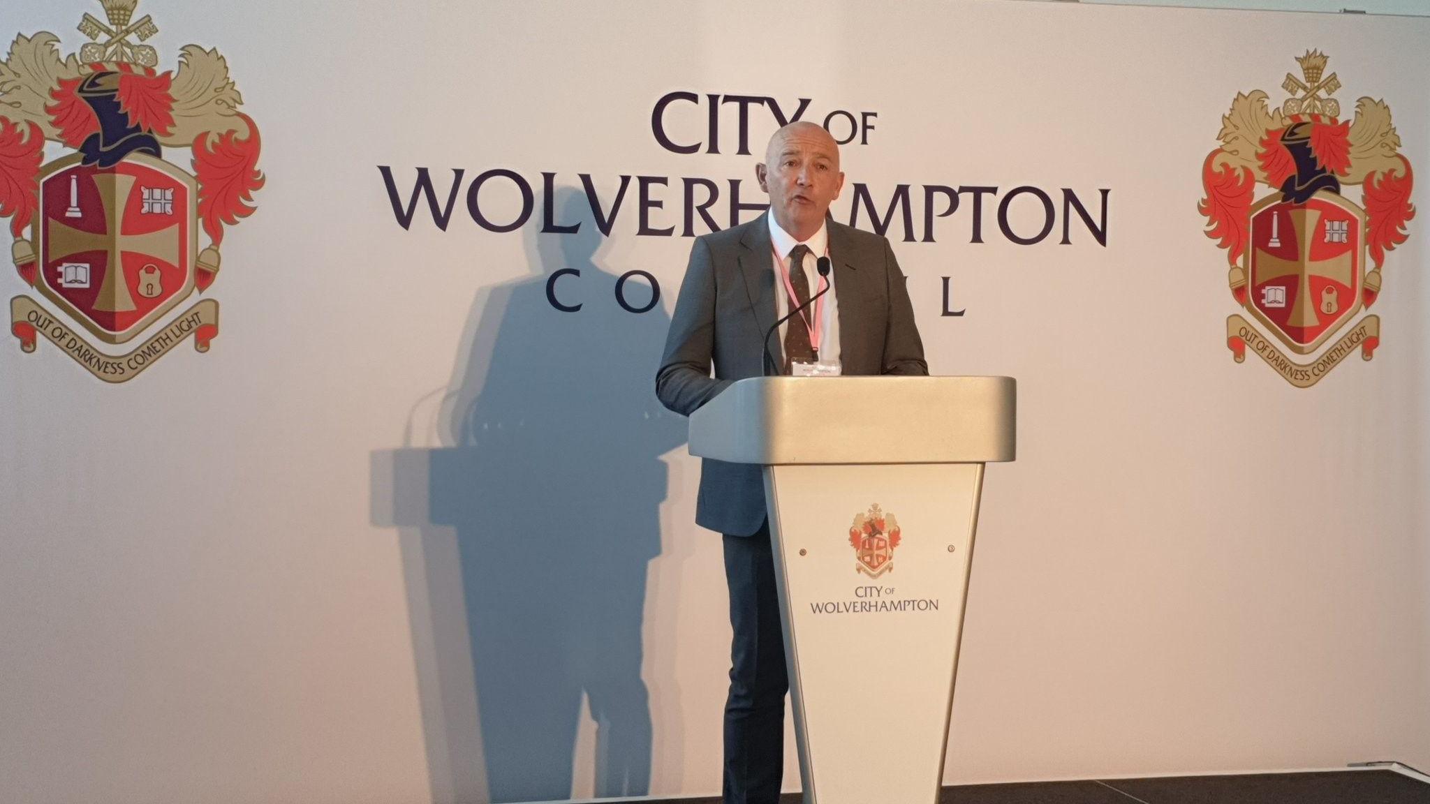 Tim Johnson is standing behind a silver plinth with the Wolverhampton council logo on it. there is a microphone which he is speaking into. He is bald and is wearing a grey suit with a white shirt. Behind him is a large white background that says "city of Wolverhampton council"