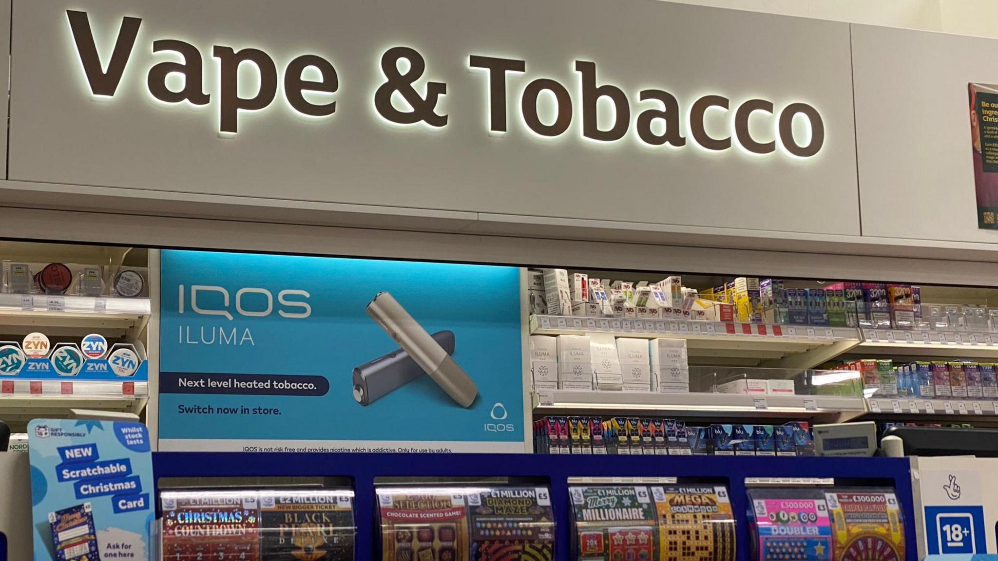 Vapes and tobacco kiosk in a Sainsbury's store in London. Sign reads 'vape and tobacco'. Below is a blue sign advertising iQos Iluma, "Next level heated tobacco", with the words "Switch now in store". 