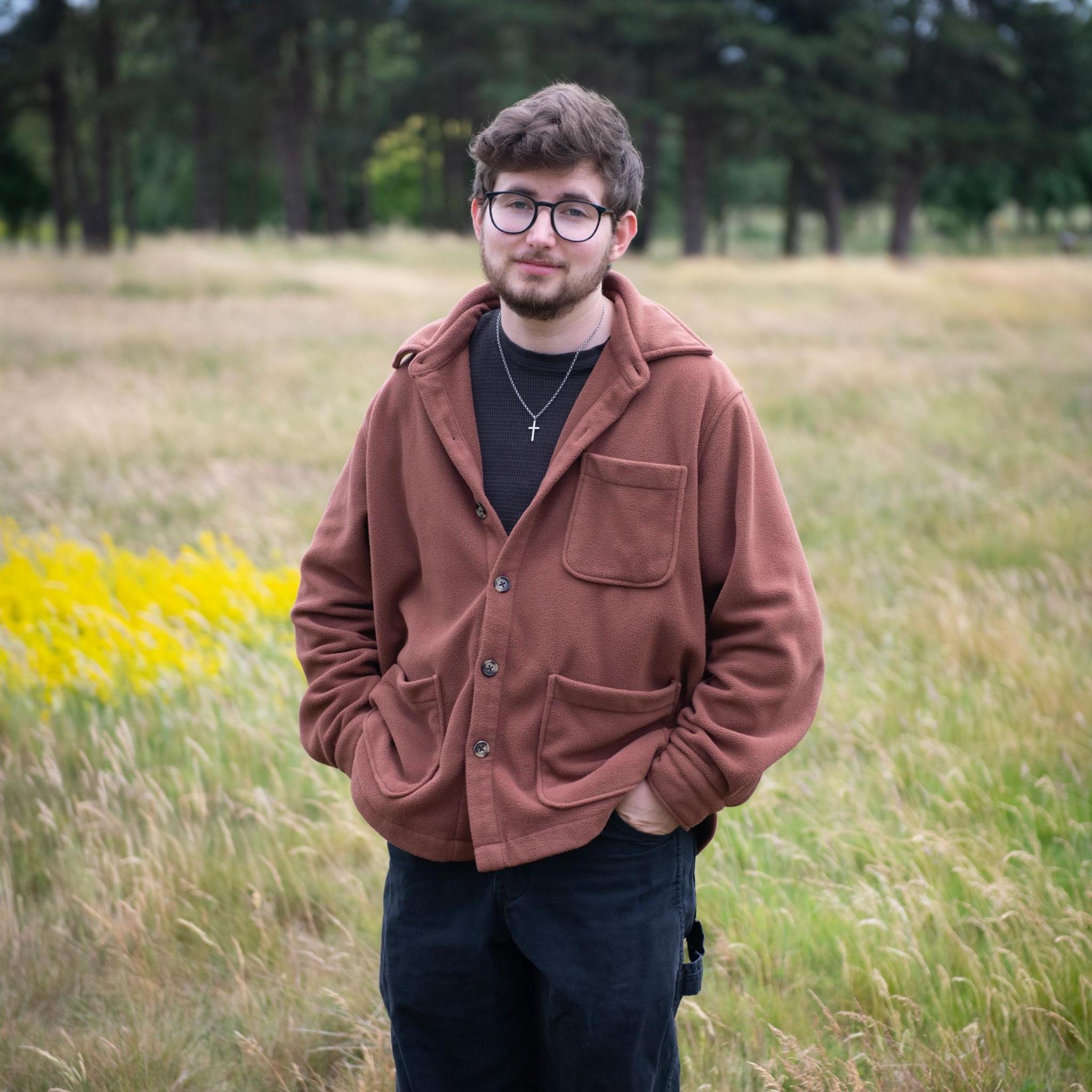 A man in a brown jacket with black trousers stands with his hands in his pockets with a long green meadow and trees in the background.