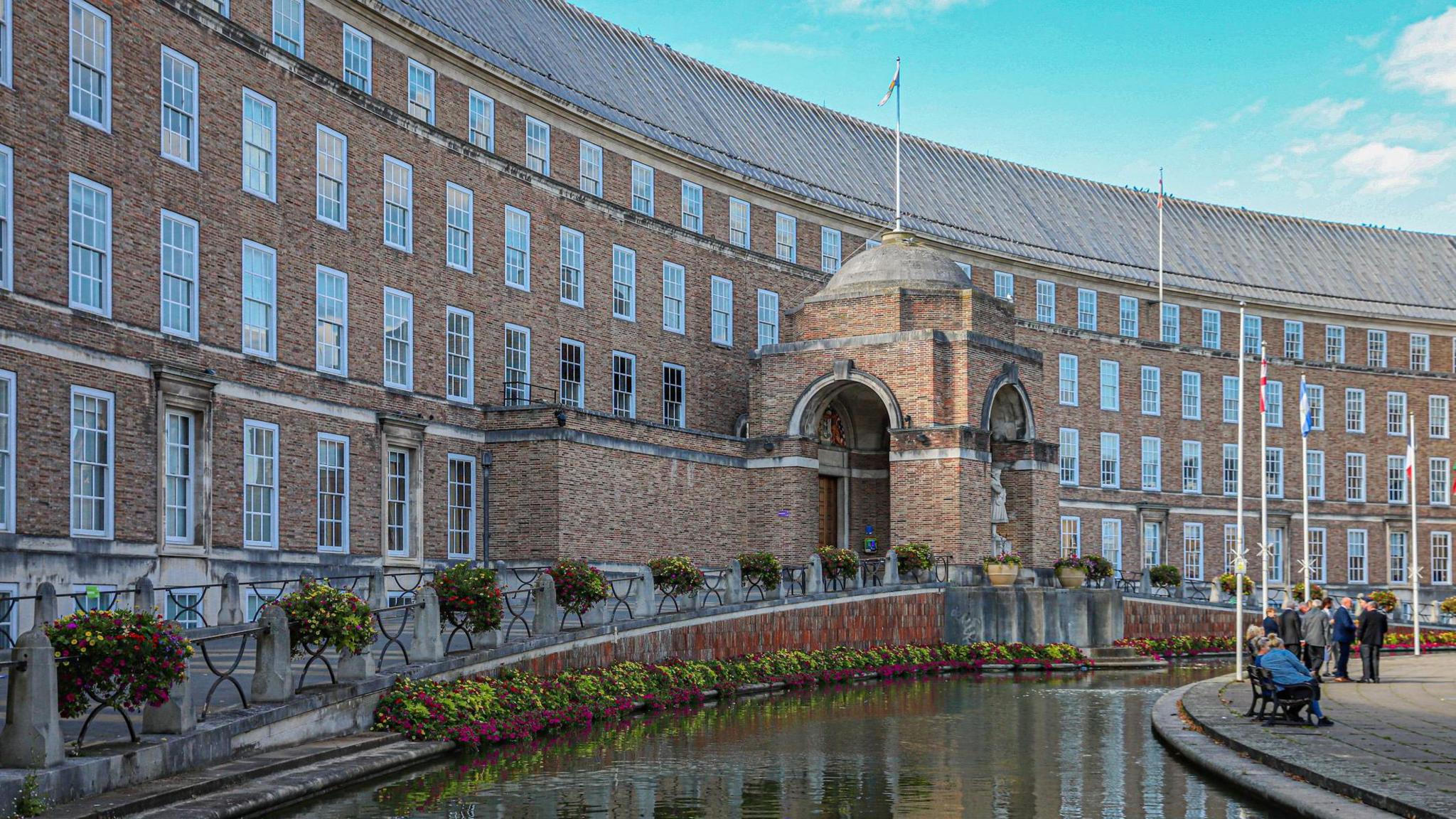 The Bristol City Council building from the outside. It is a crescent-shaped red brick building with rows of windows. There is a water feature in the front of the building and some flag poles.