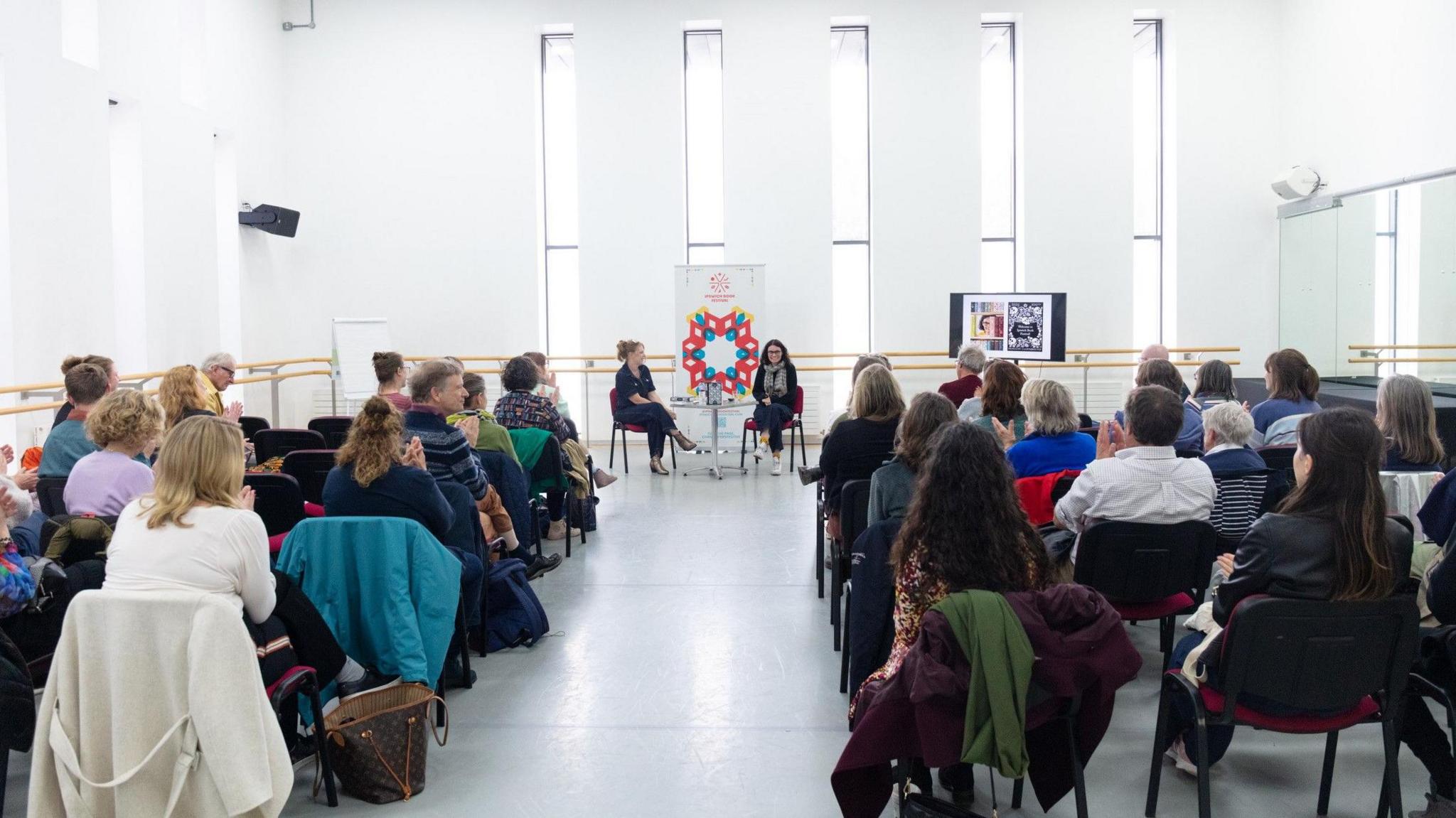 A large white room is filled with people occupying seats that face two women sitting at the front. Some of the people are clapping.