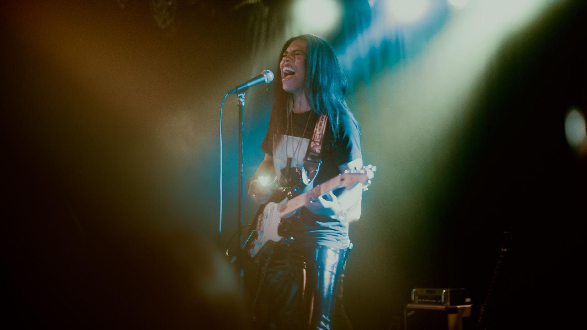 A man with long black hair singing into a microphone and playing a guitar. He's wearing leather trousers and a black top and lights are shining down on him