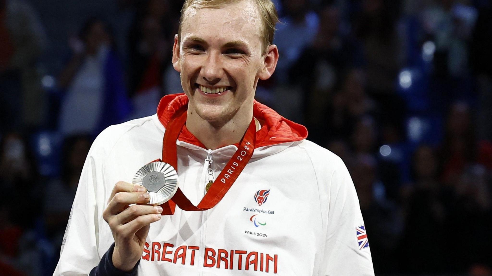 Piers Gilliver smiles as he holds a silver medal up at the Paris Paralympics. He is wearing a white top with Great Britain written on it in red letterd