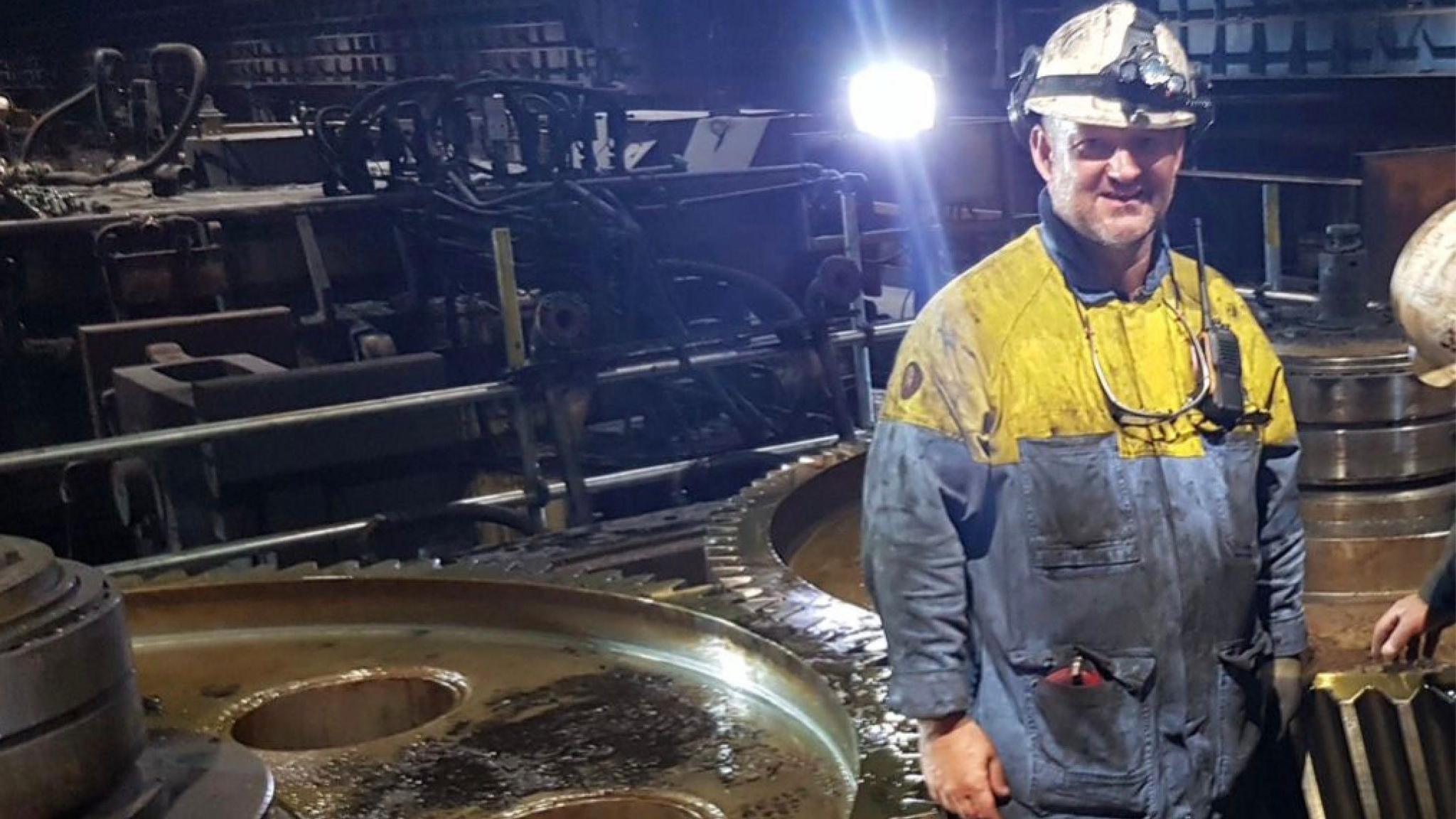 Man wearing blue and yellow overalls in a white helmet standing to the right of frame with industrial sized cogs behind him and a bright, white light in the background. He also has a pair of protective goggles hanging around his chest and a walkie-talkie attached to the left side of his chest. You can also see the hand and the white heltmet of another worker peaking into the right side of the frame.