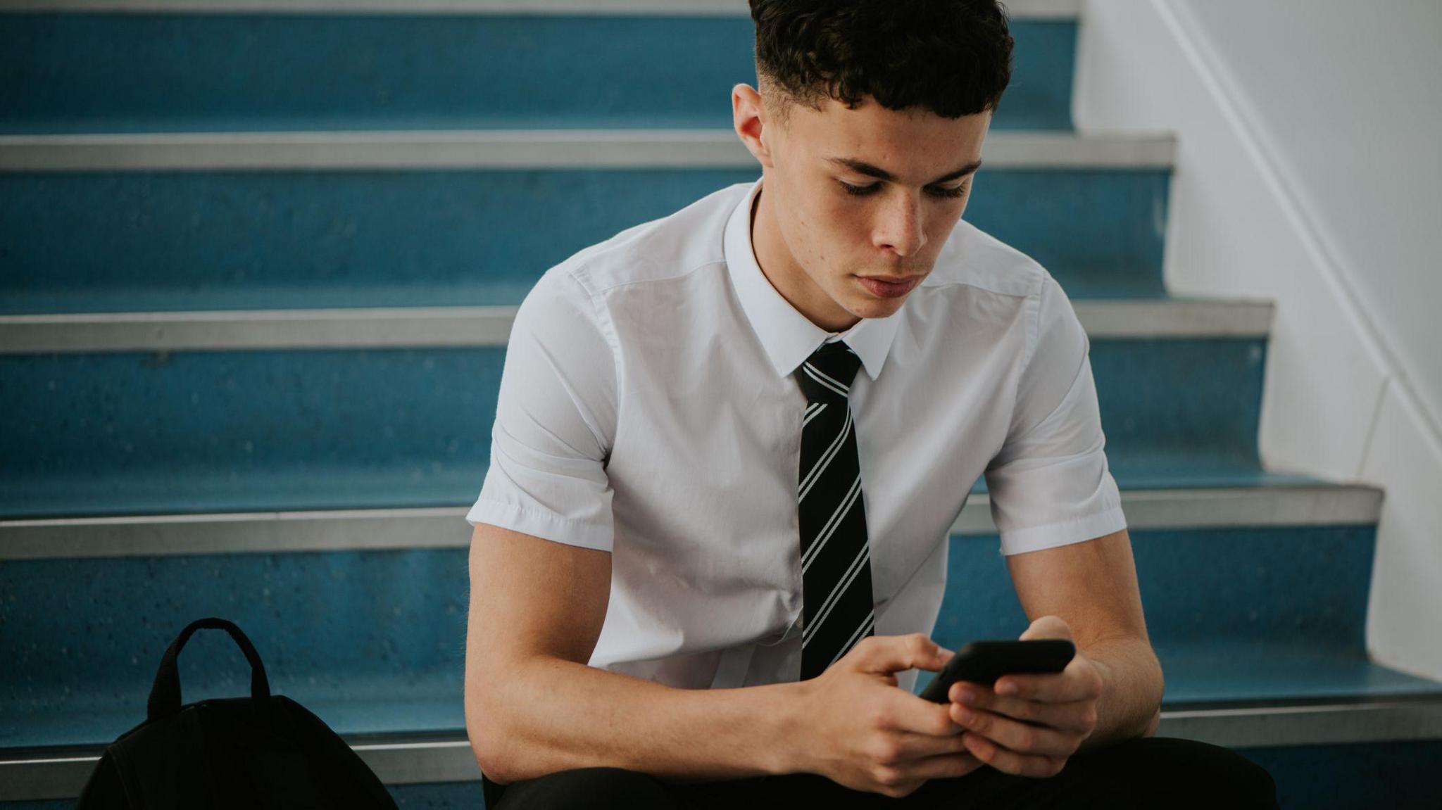 A teenage lad  successful  school, wearing a garment  and tie, sat connected  immoderate   stairs, utilizing his phone