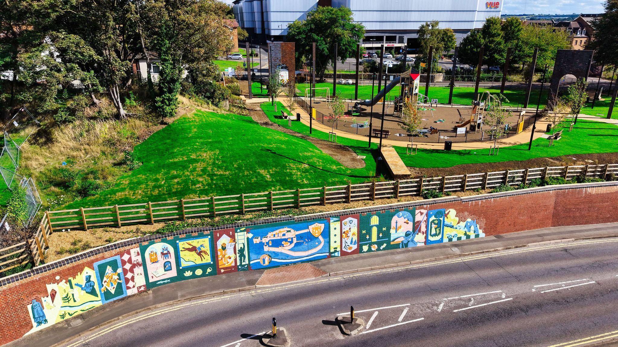 A heritage park, complete with a play area, grass, trees, wooden fence, by a cinema, and a road. A brick wall, in front of it, also features a colour mural, painted on it.