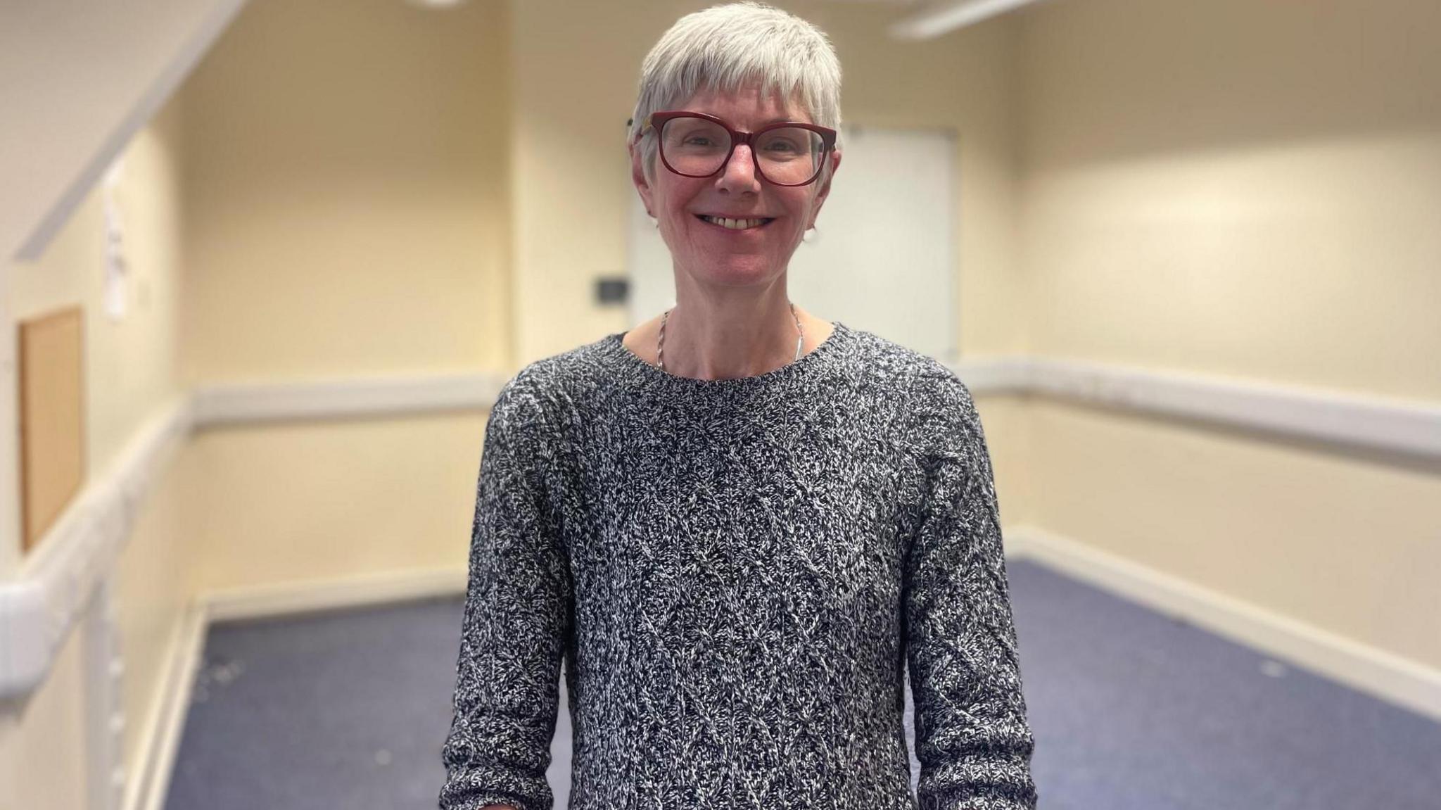 A woman with short grey hair and glasses smiles at the camera. There is a bare room behind her