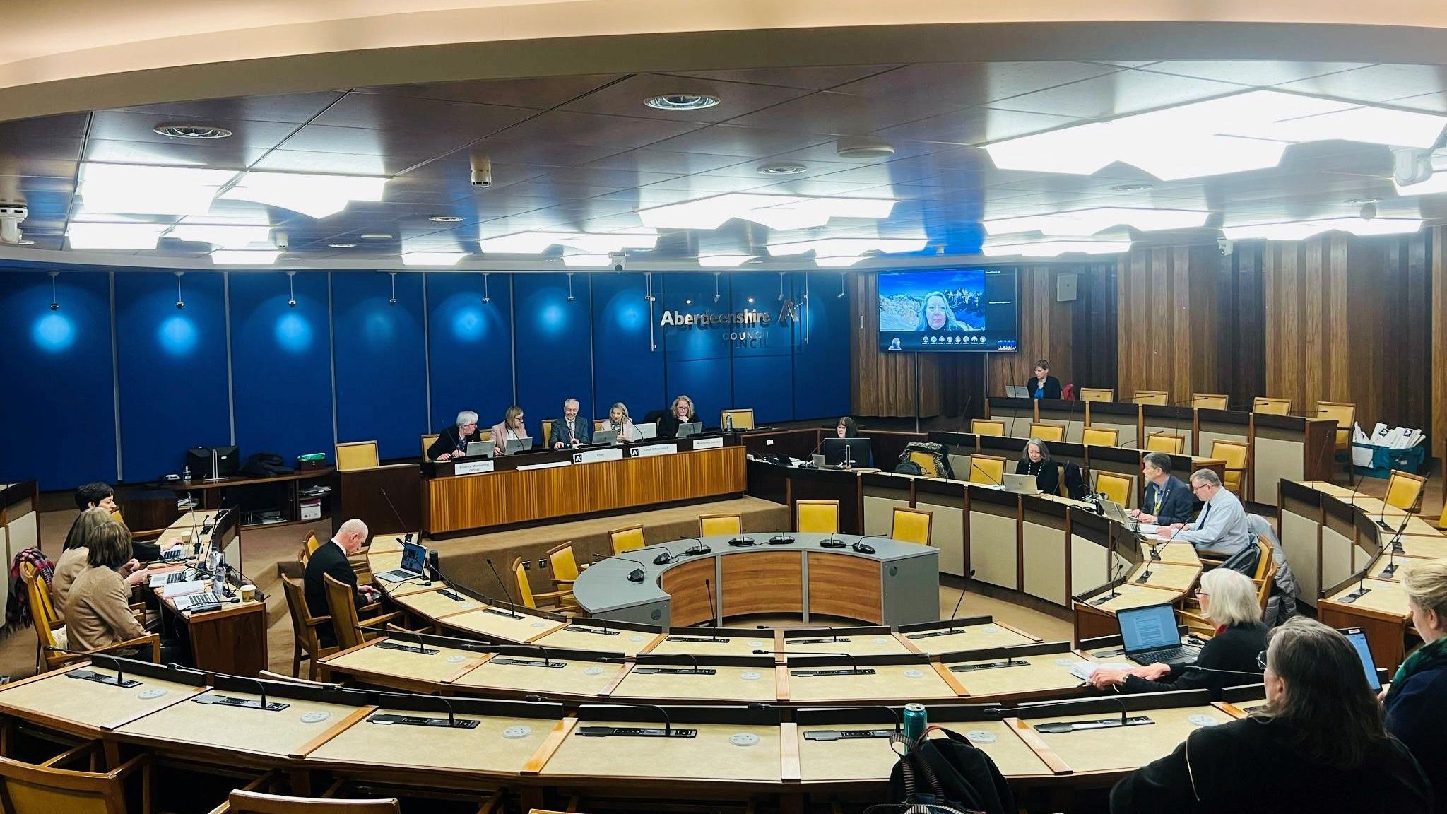 A meeting of the Integration Joint Board (IJB) - the body which runs health and social care services in the region - with people sitting in a meeting room, under bright lights, with an Aberdeenshire Council sign on wall.